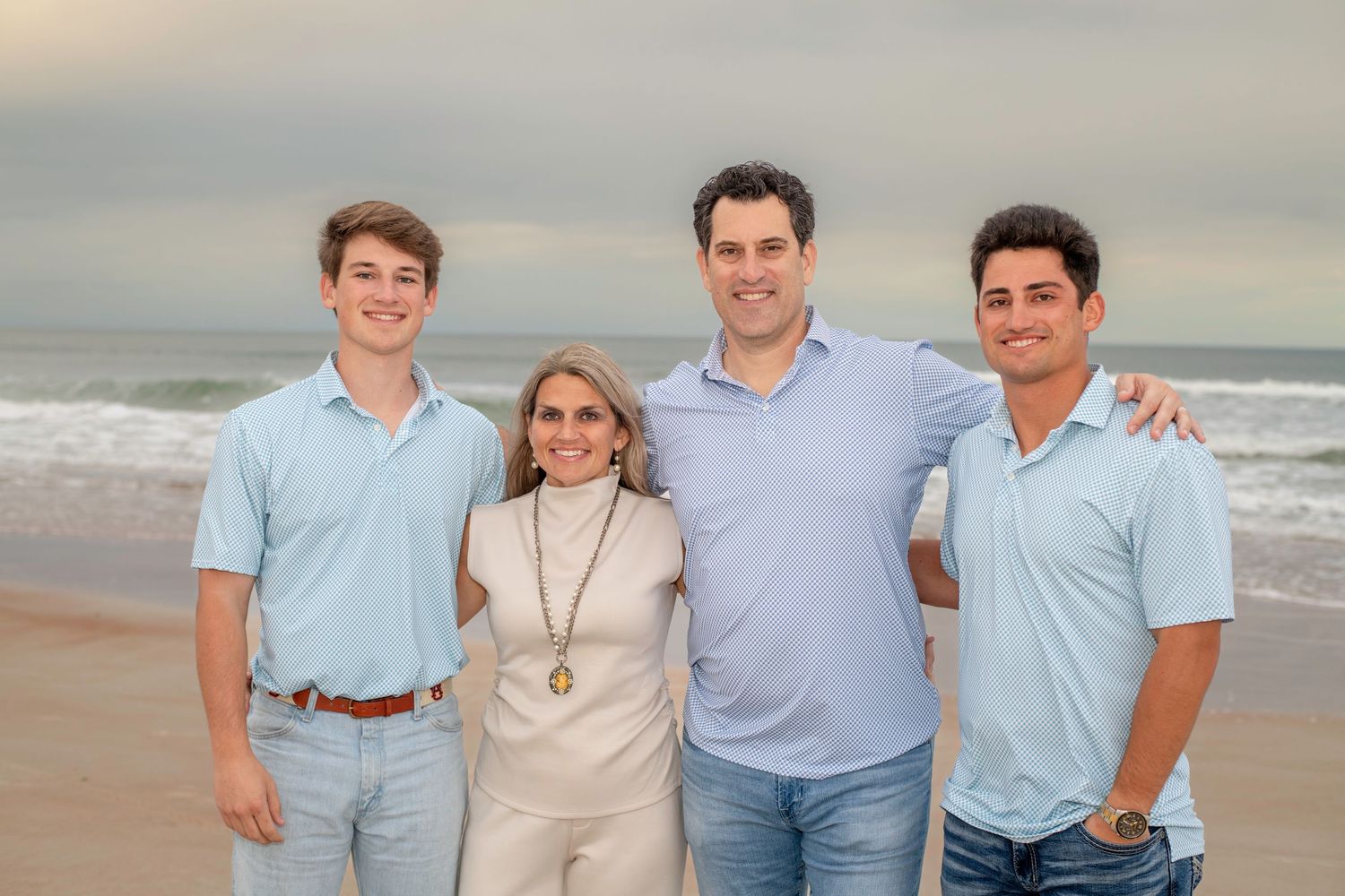 Family of four smiling together on a beach with ocean waves in the background on a cloudy day.
