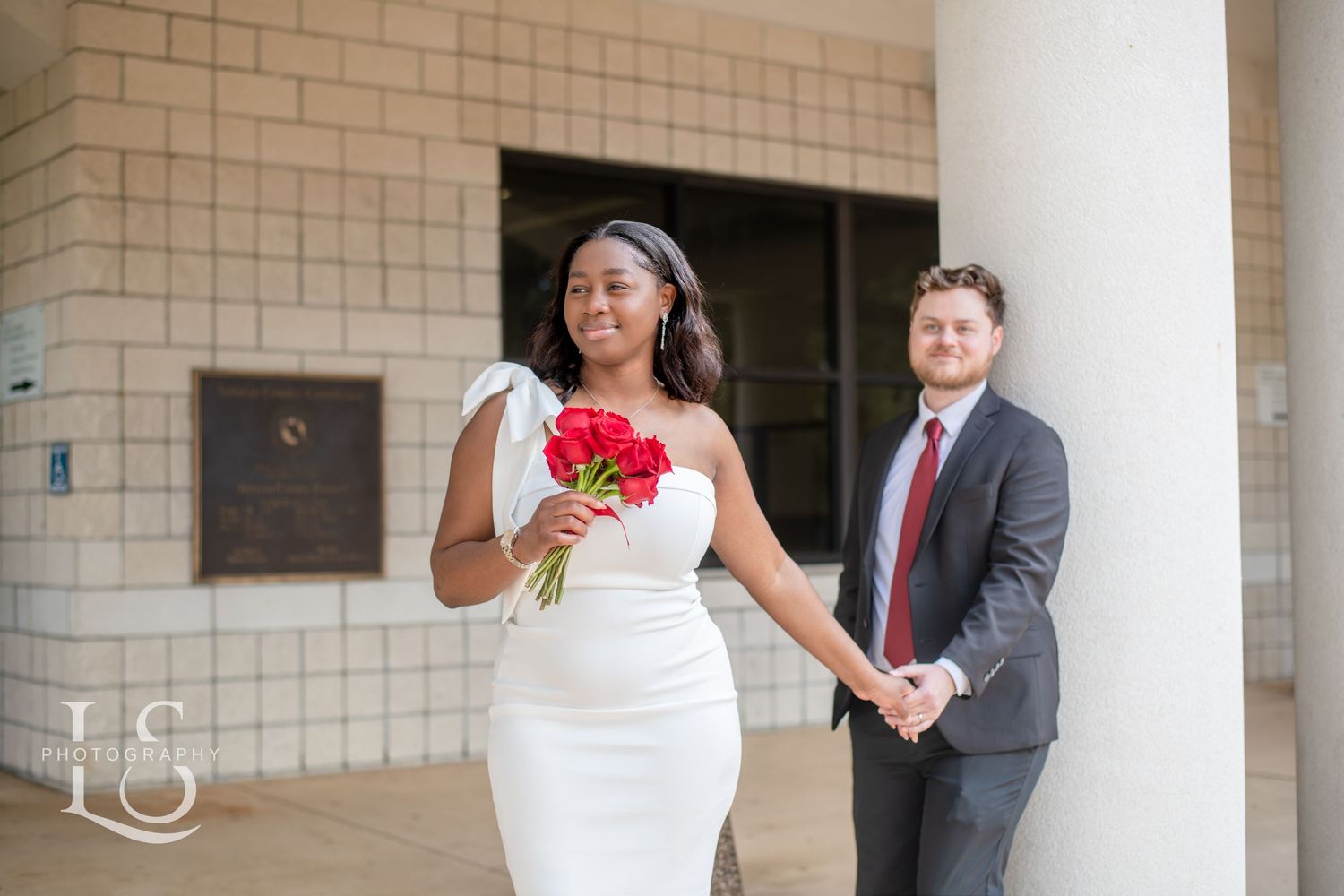 A couple poses in formal wedding attire with red roses against a brick and column backdrop.