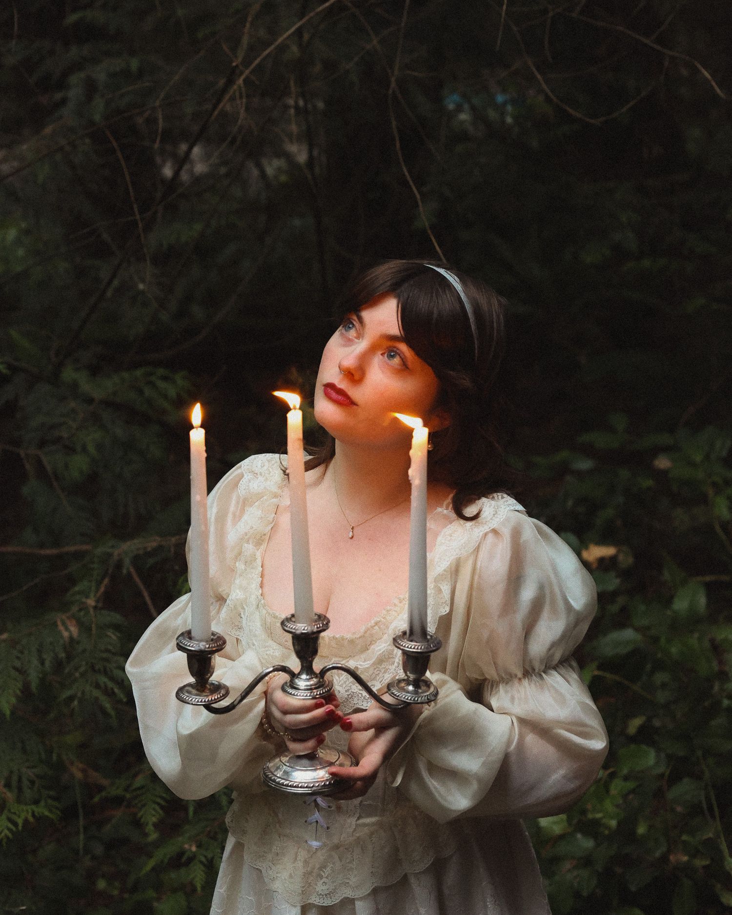 A model wearing a flow white dress holding a lit candelabra walks in the forest.