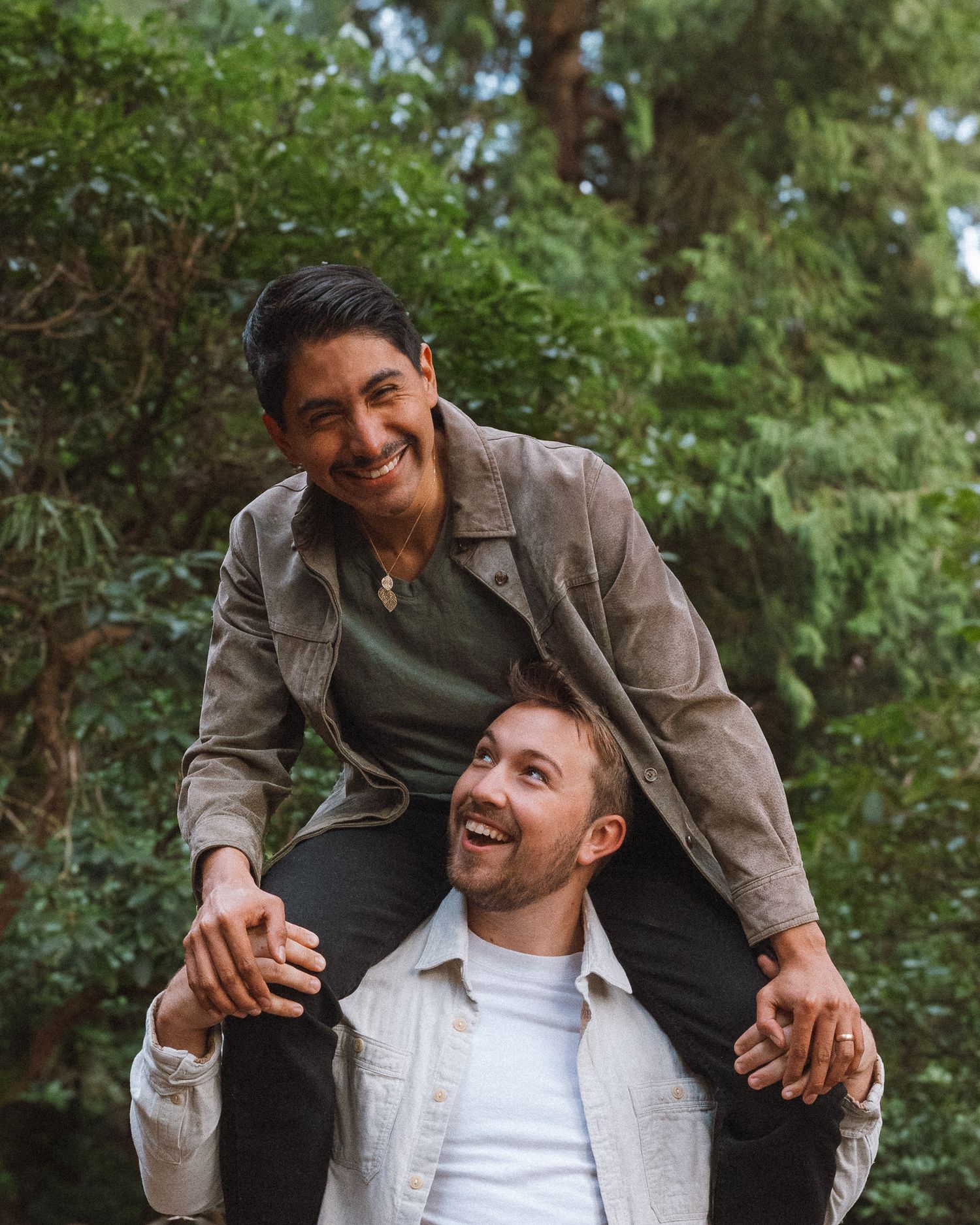 A couple in a forest smiles and laughs at each other during a photoshoot.