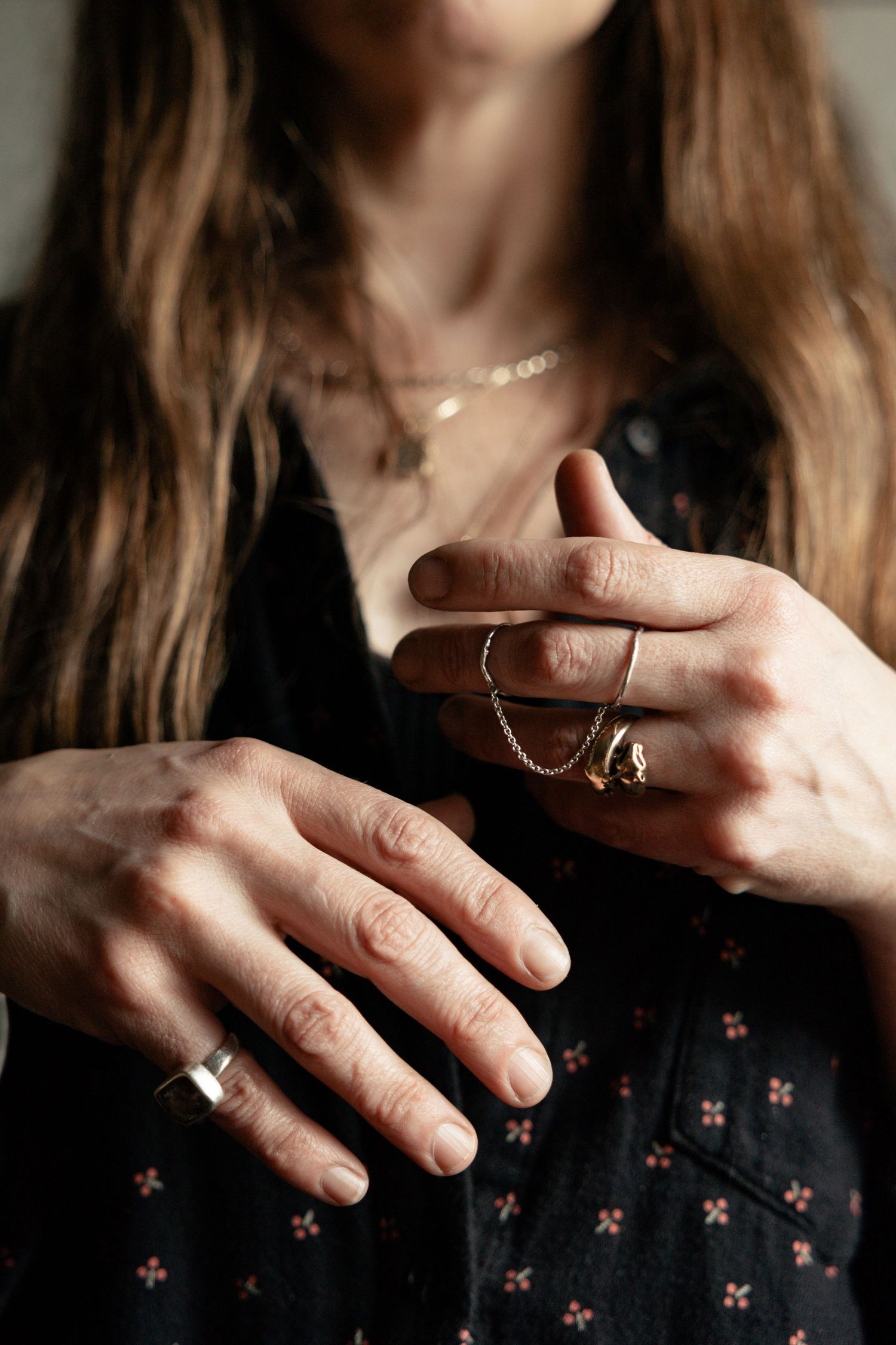 Close-up of hands wearing delicate jewelry and rings against a dark patterned fabric background.