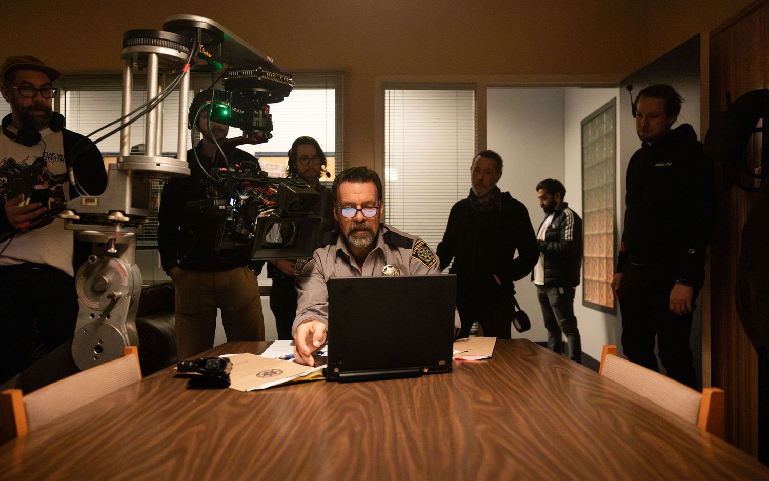 Behind the scenes shot showing a camera crew filming a scene with a person working at a laptop in an office setting.
