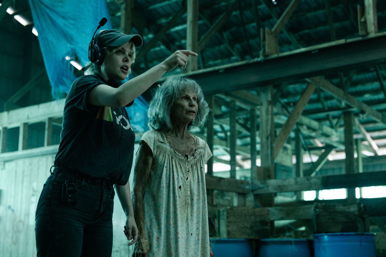 Two figures stand in a dark industrial setting with teal lighting, one pointing while the other wears a white nightgown.