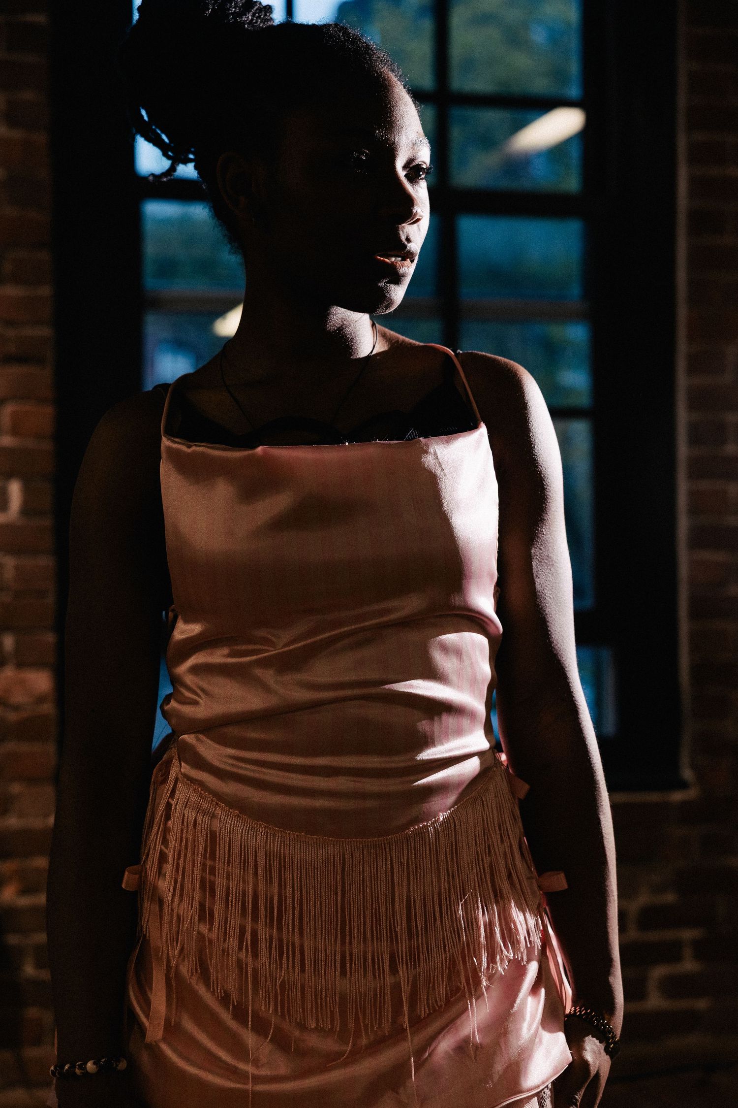 An artistic portrait shows a person in a pink satin dress with fringe detail standing against a dark brick wall.
