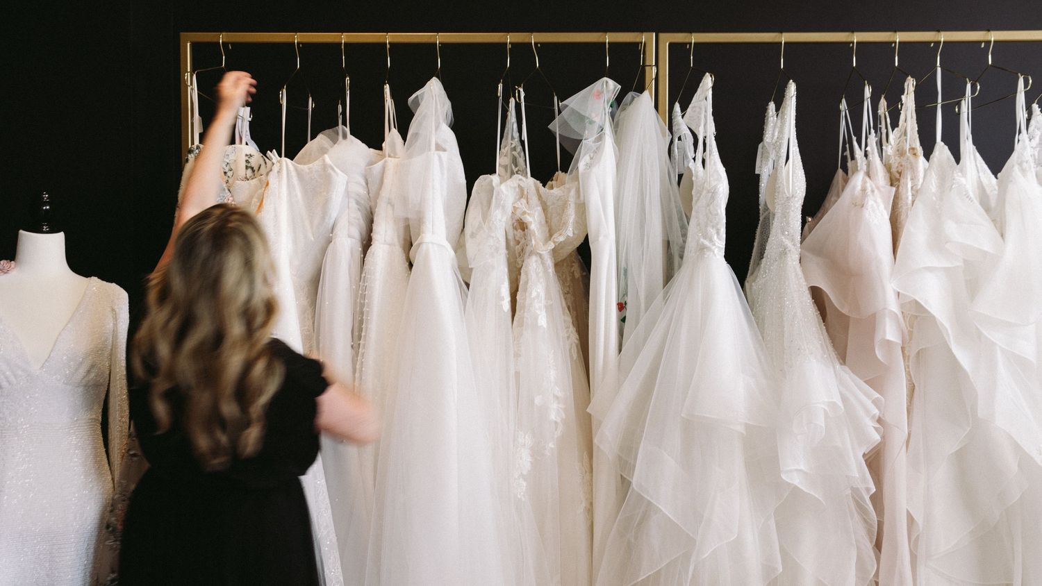 A rack of white wedding dresses hanging in a bridal boutique with someone browsing the selection.