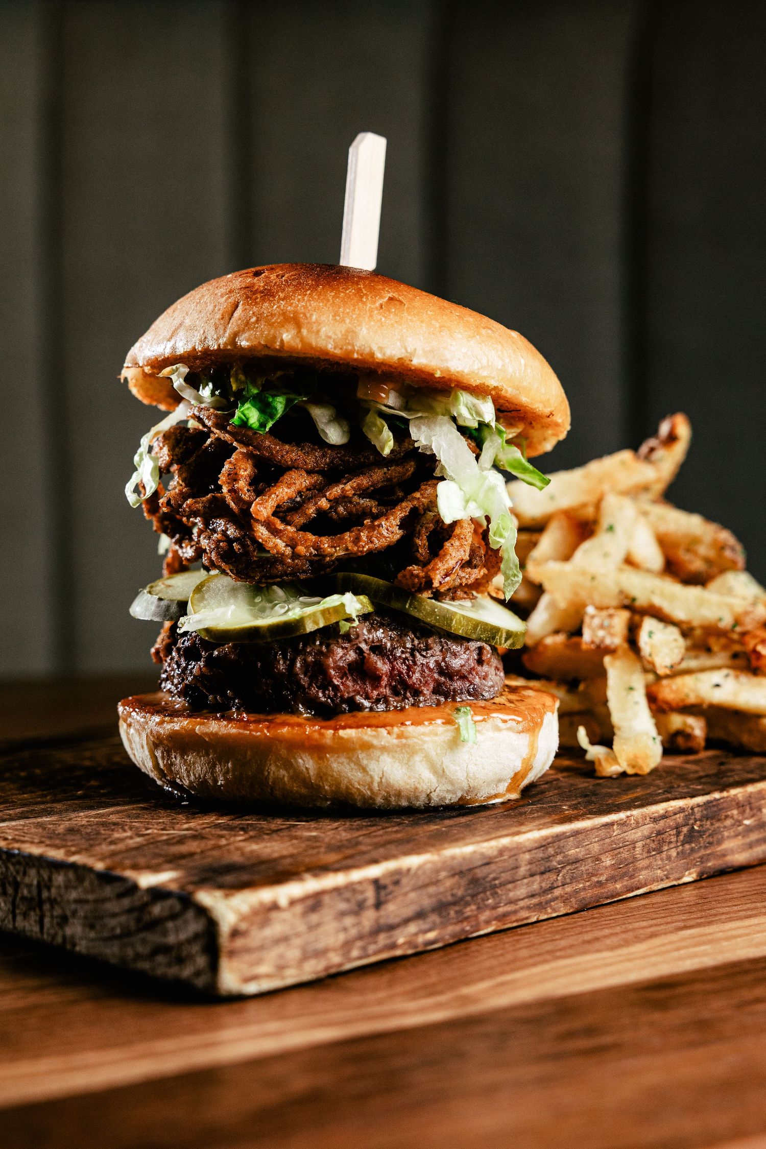 Gourmet burger stacked with crispy onions and toppings served with hand-cut fries on wooden board.