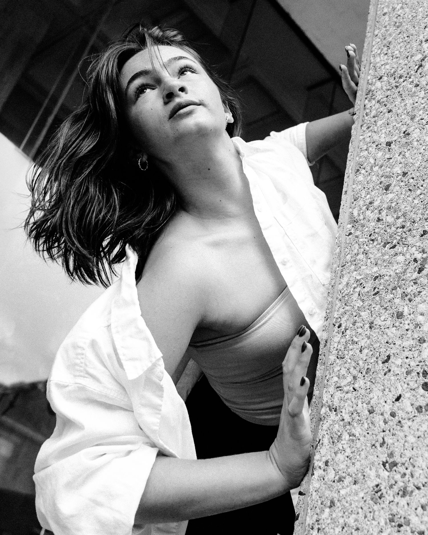 Artistic black and white portrait shot leaning against stone pillar wearing white shirt and tank top.
