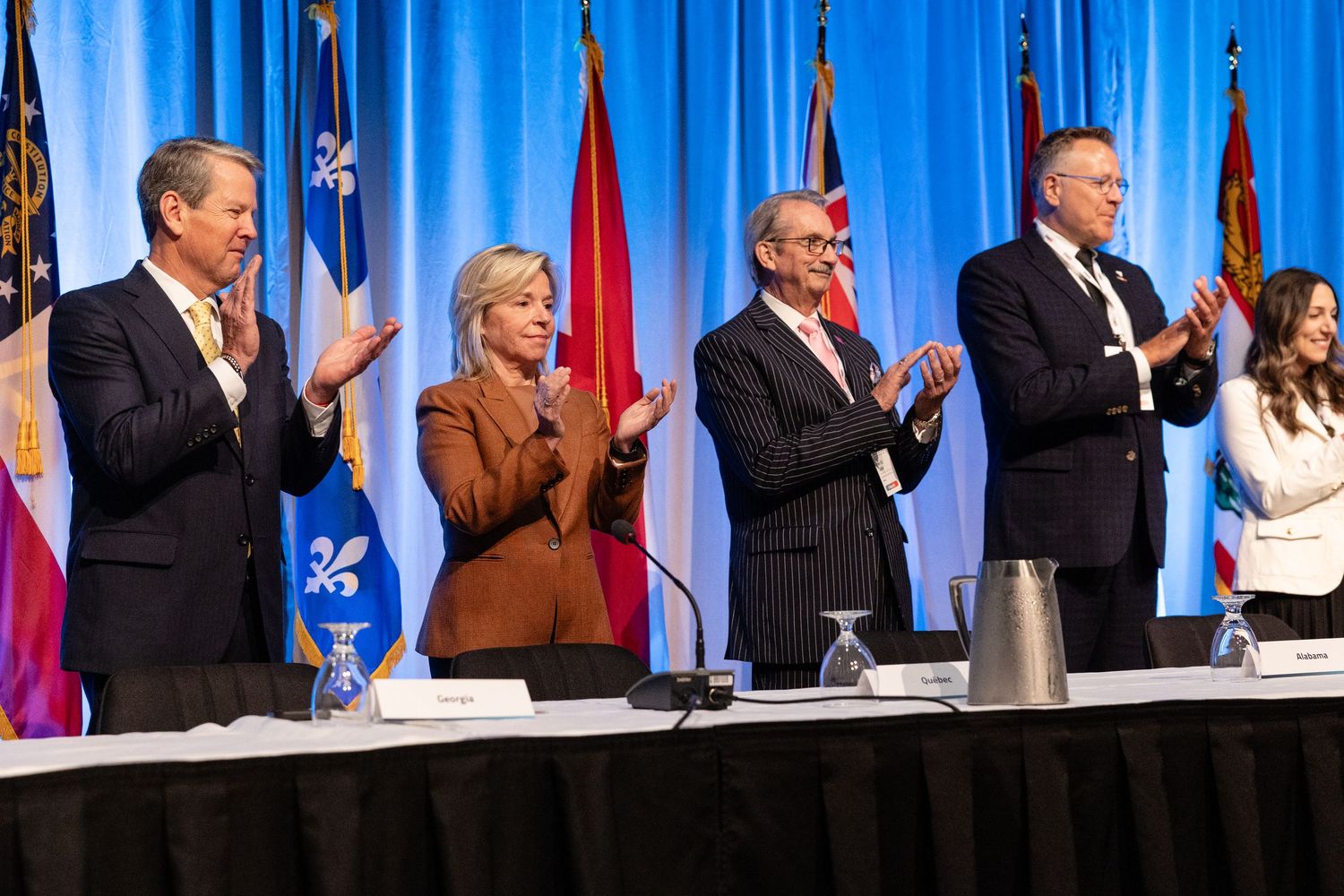 Political figures applaud while standing at a podium with flags and blue curtains behind them during a formal event.