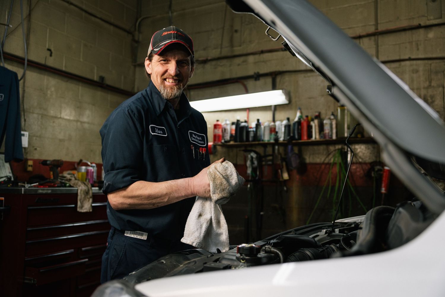 Auto mechanic in blue uniform holds a shop rag while working on a car engine in a repair garage.