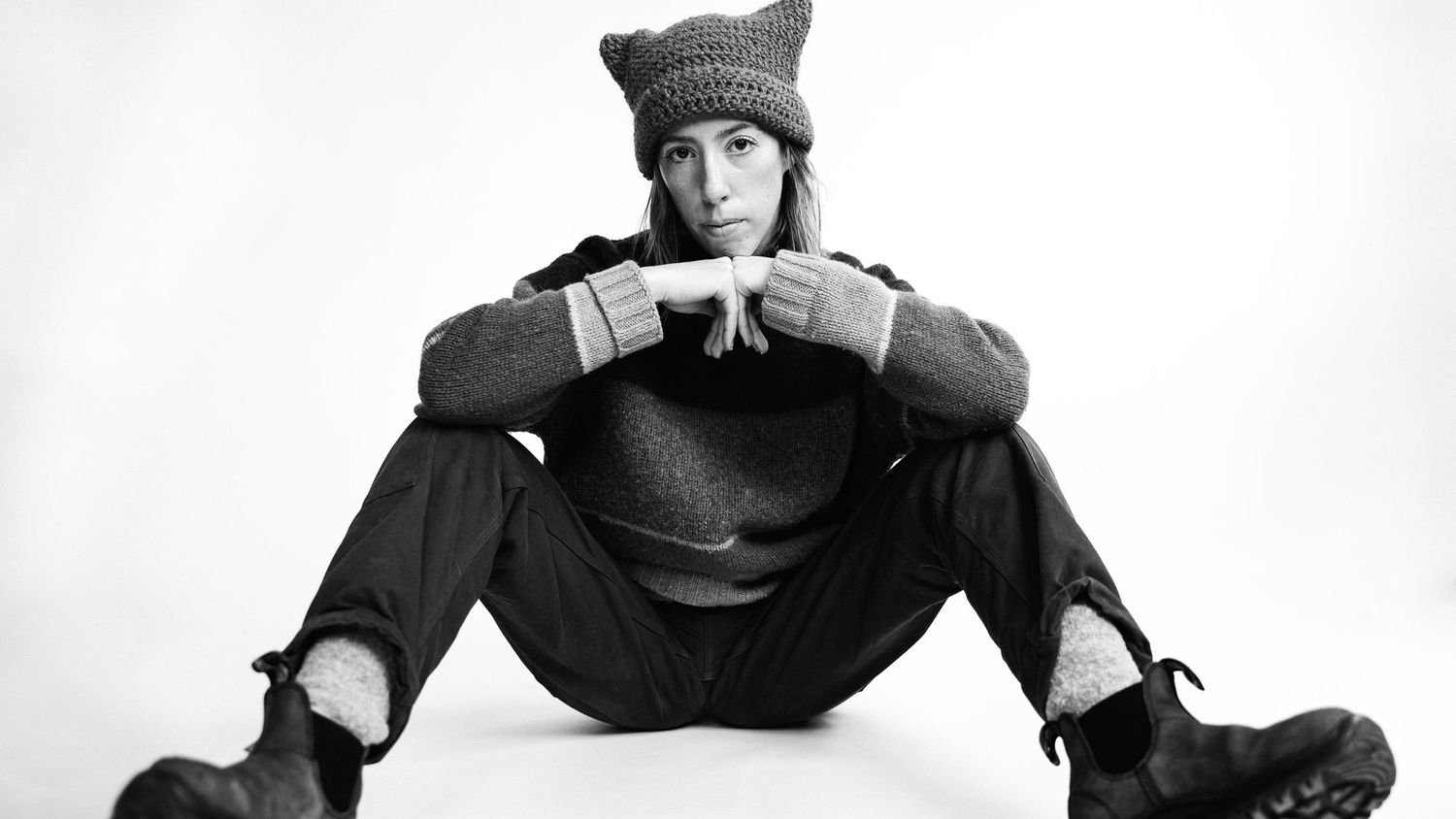 A figure in a knit hat and dark clothing sits in a casual pose against a white background.