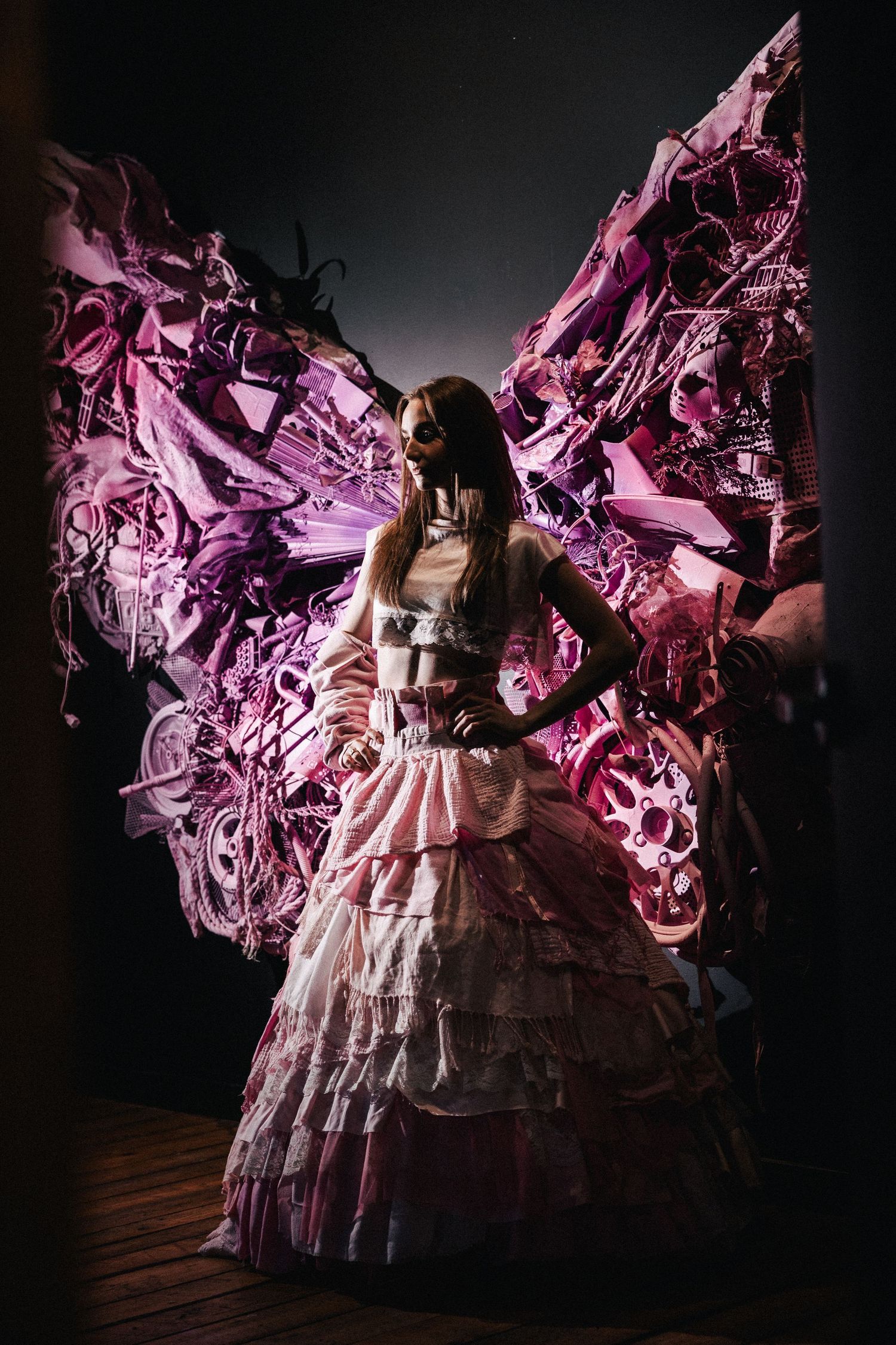 Dark fantasy woman in ruffled pink dress with illuminated glowing butterfly wings against black background.