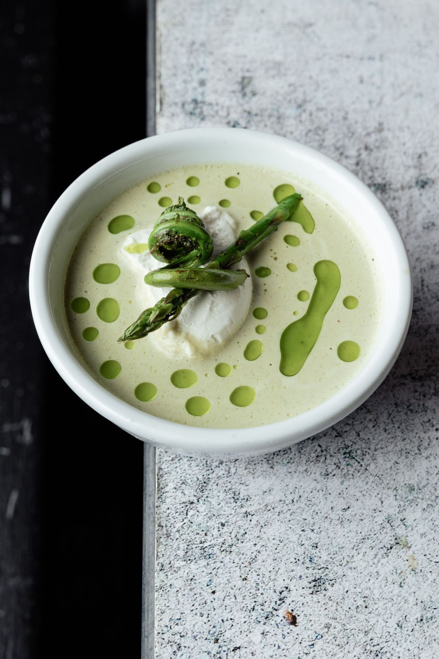 Elegant white asparagus soup garnished with fresh asparagus spears and olive oil dots in a white bowl on concrete surface.