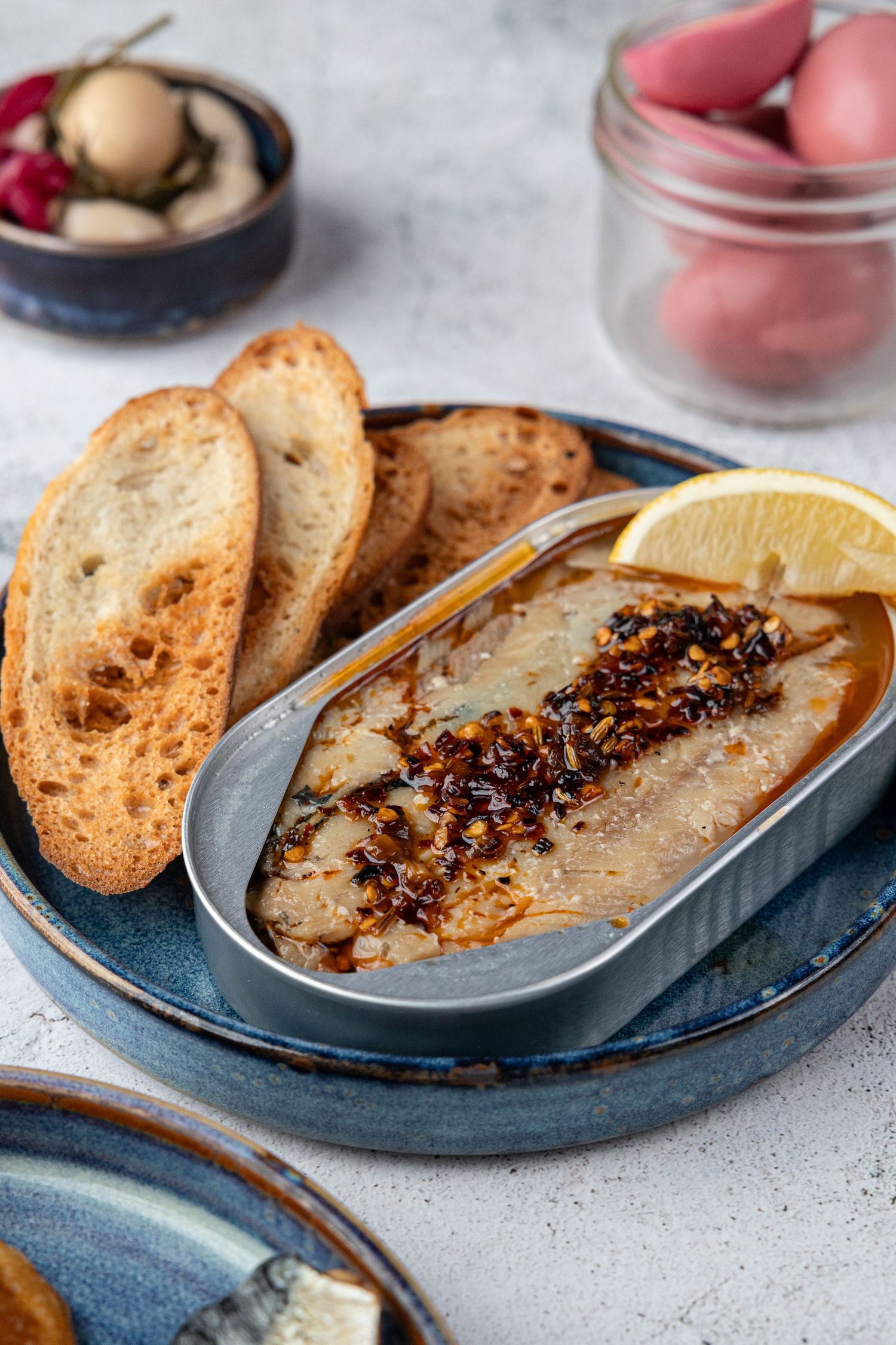 Grilled sardines with crispy bread slices served on a blue ceramic plate with lemon wedge and chili oil garnish.