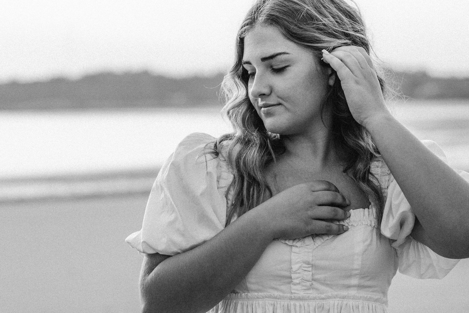 Black and white portrait of person with long wavy hair wearing white dress looking down pensively at beach.