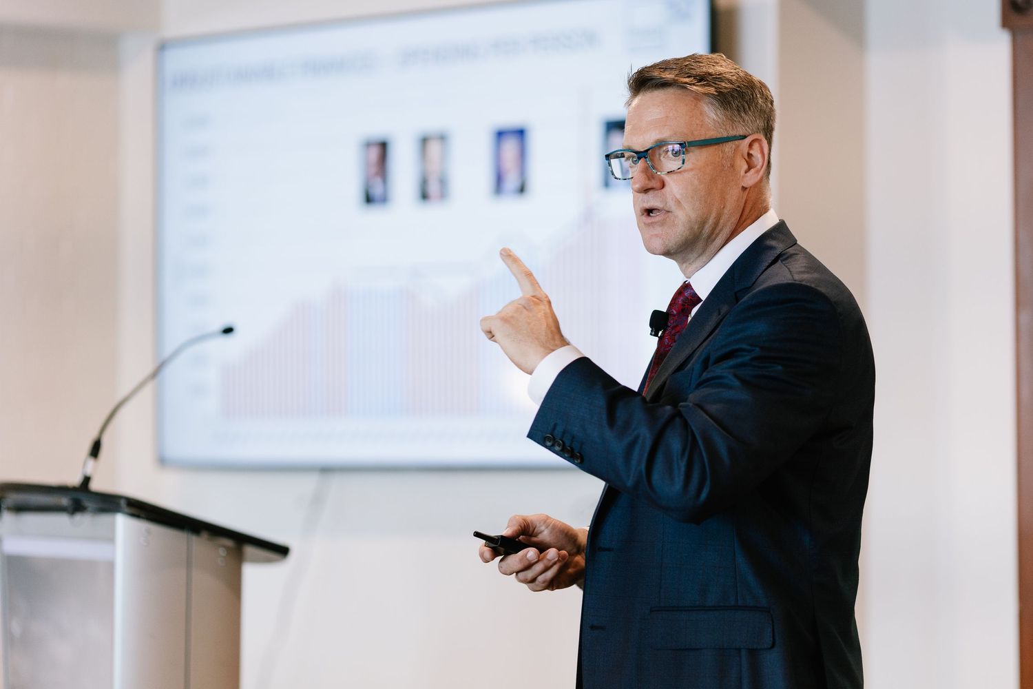 A presenter in a navy suit and red tie gestures while speaking at a business conference podium.