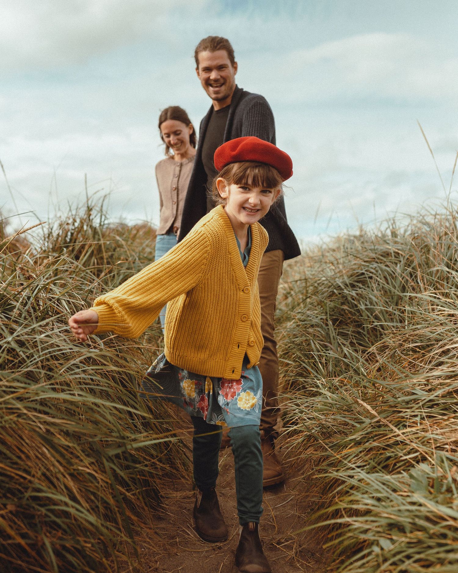 Family playfully walks through tall grass and rushes at sunset along a coastal pathway.