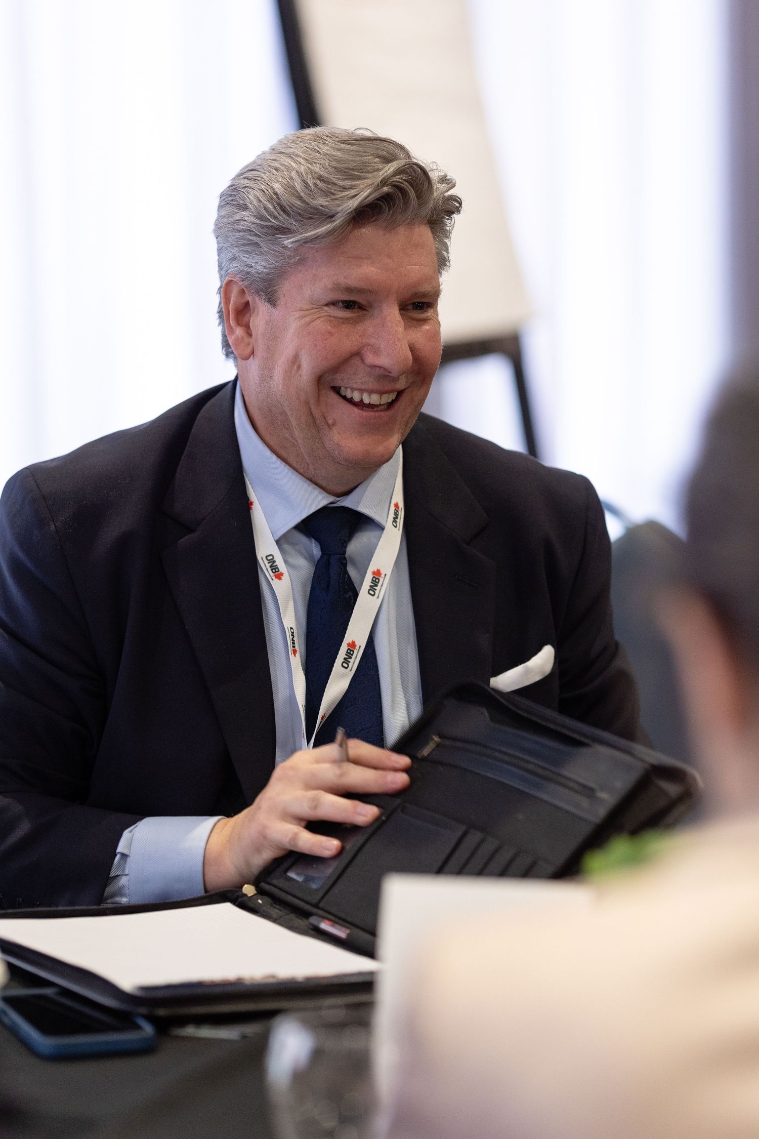 A businessman in a dark suit laughs during a meeting while sitting at a conference table.
