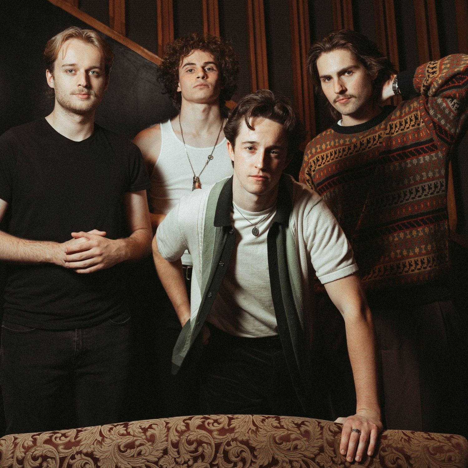 A moody portrait of a four-member band posing together against dark decor and patterned upholstery.