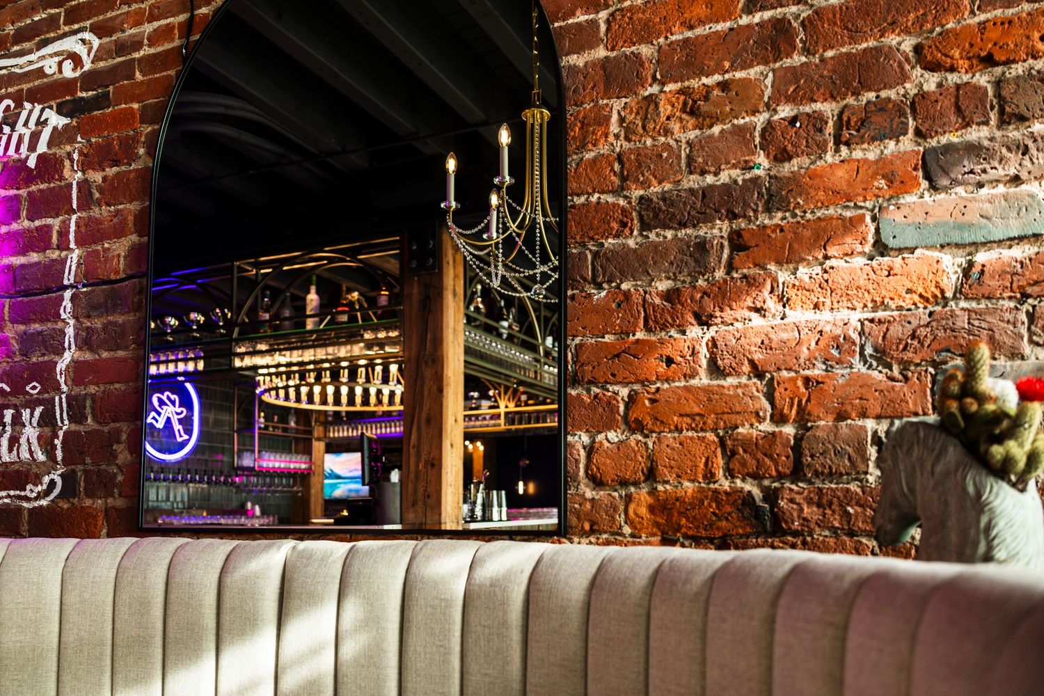 Exposed brick wall and plush beige booth seating in a dimly lit bar with bottles displayed through an archway.