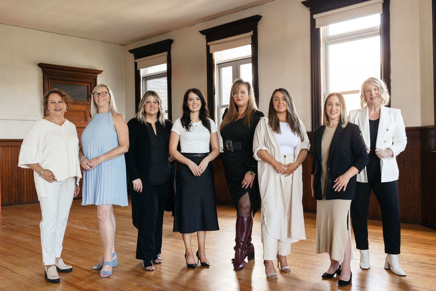 Professional photo of a business team standing together in formal attire on a hardwood floor with tall windows in background.