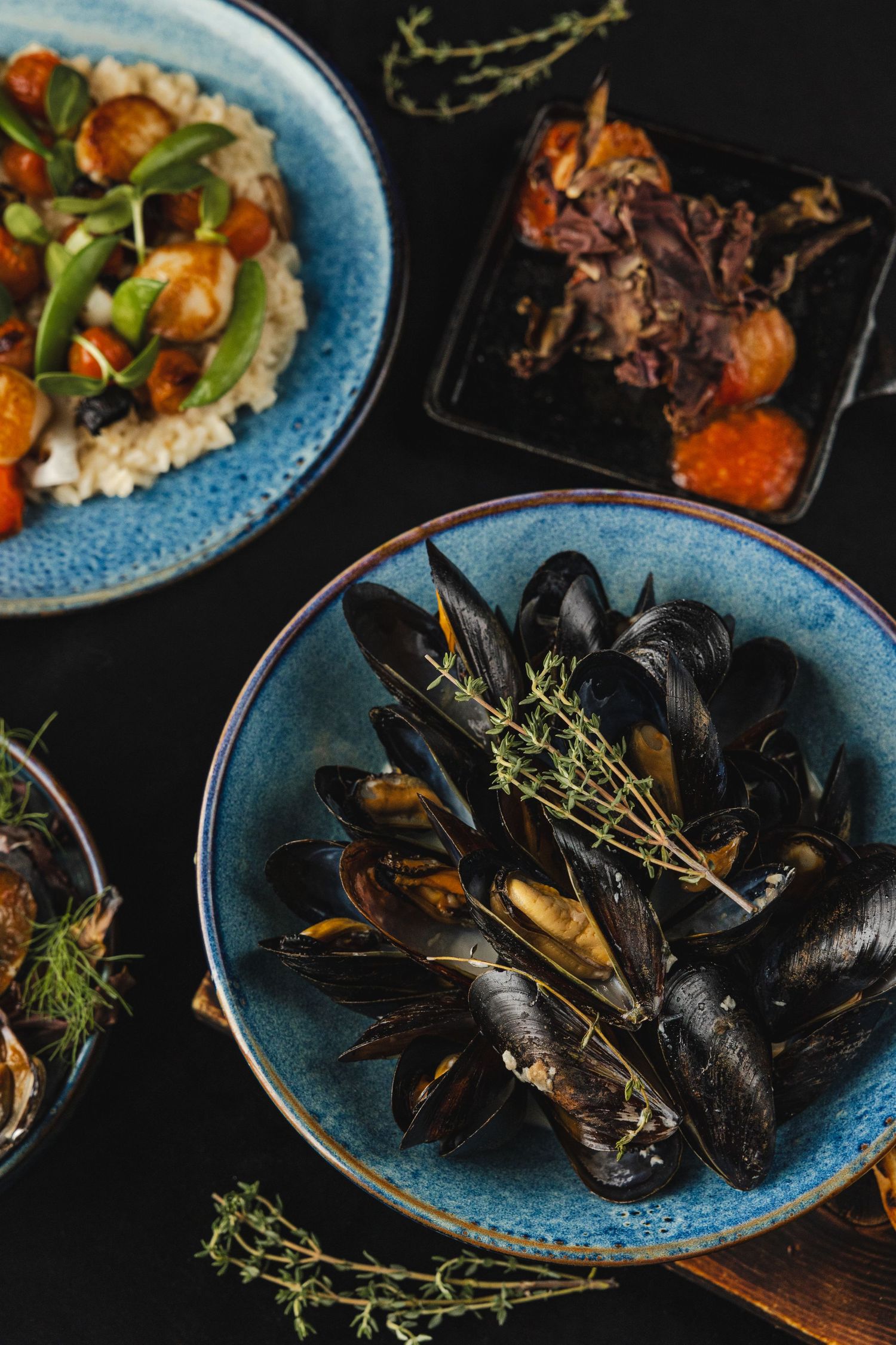 Fresh mussels garnished with herbs served on blue ceramic plates alongside other seafood dishes on a dark table.