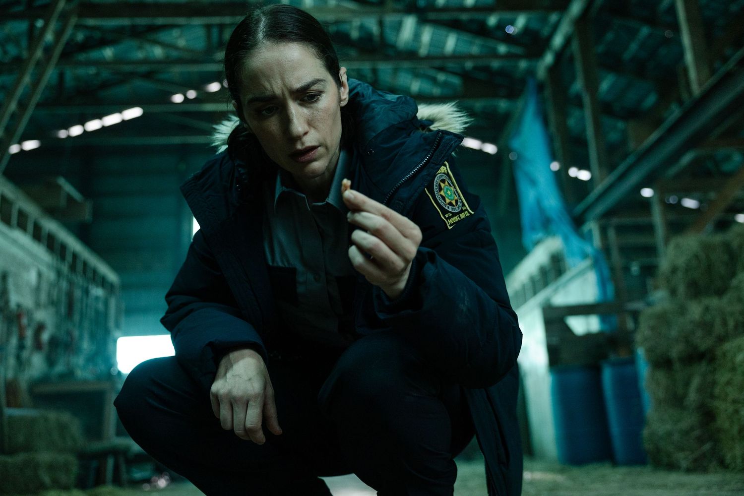 Figure in dark police uniform crouches in dimly lit industrial setting with blue-green lighting.