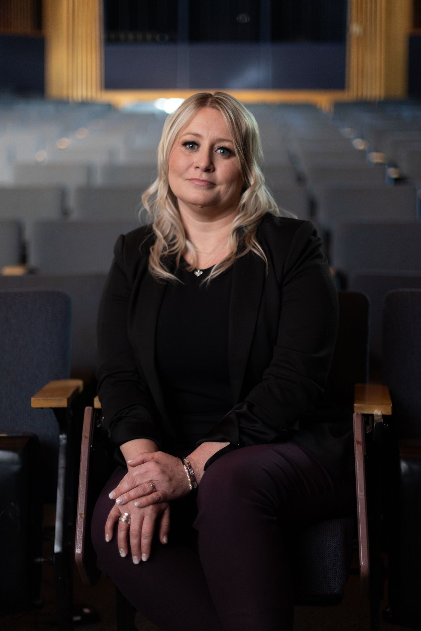 Professional portrait of someone sitting in an auditorium seat wearing a black blazer with blonde hair.