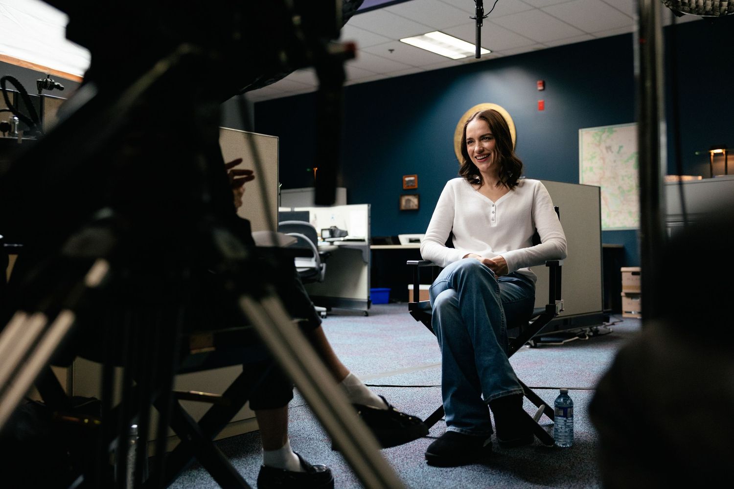 Someone in white headphones and sweater sits in a dimly lit office space with camera equipment visible in foreground.
