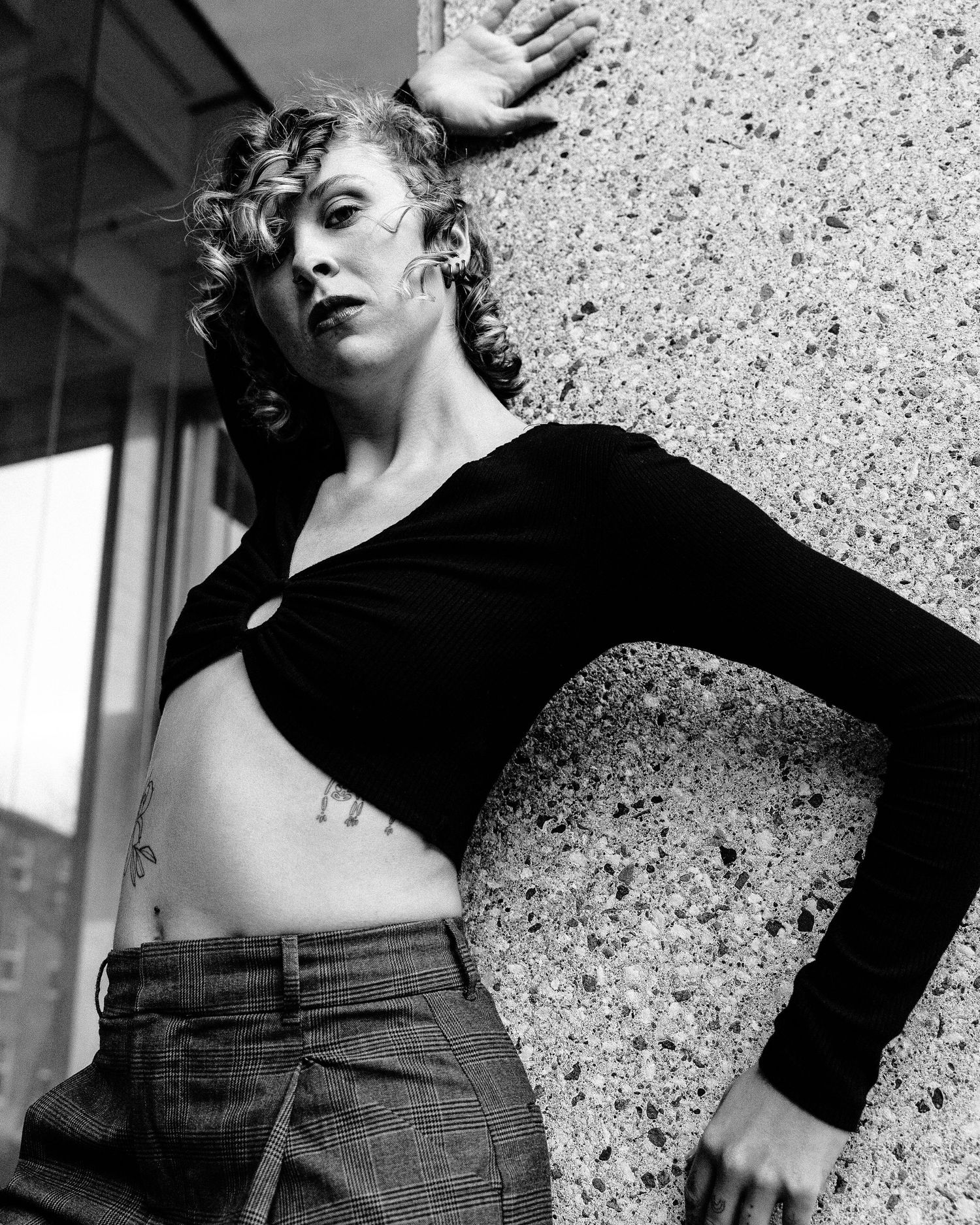 A dramatic black and white portrait shot against a textured wall shows a figure in black long sleeves and high-waisted pants.