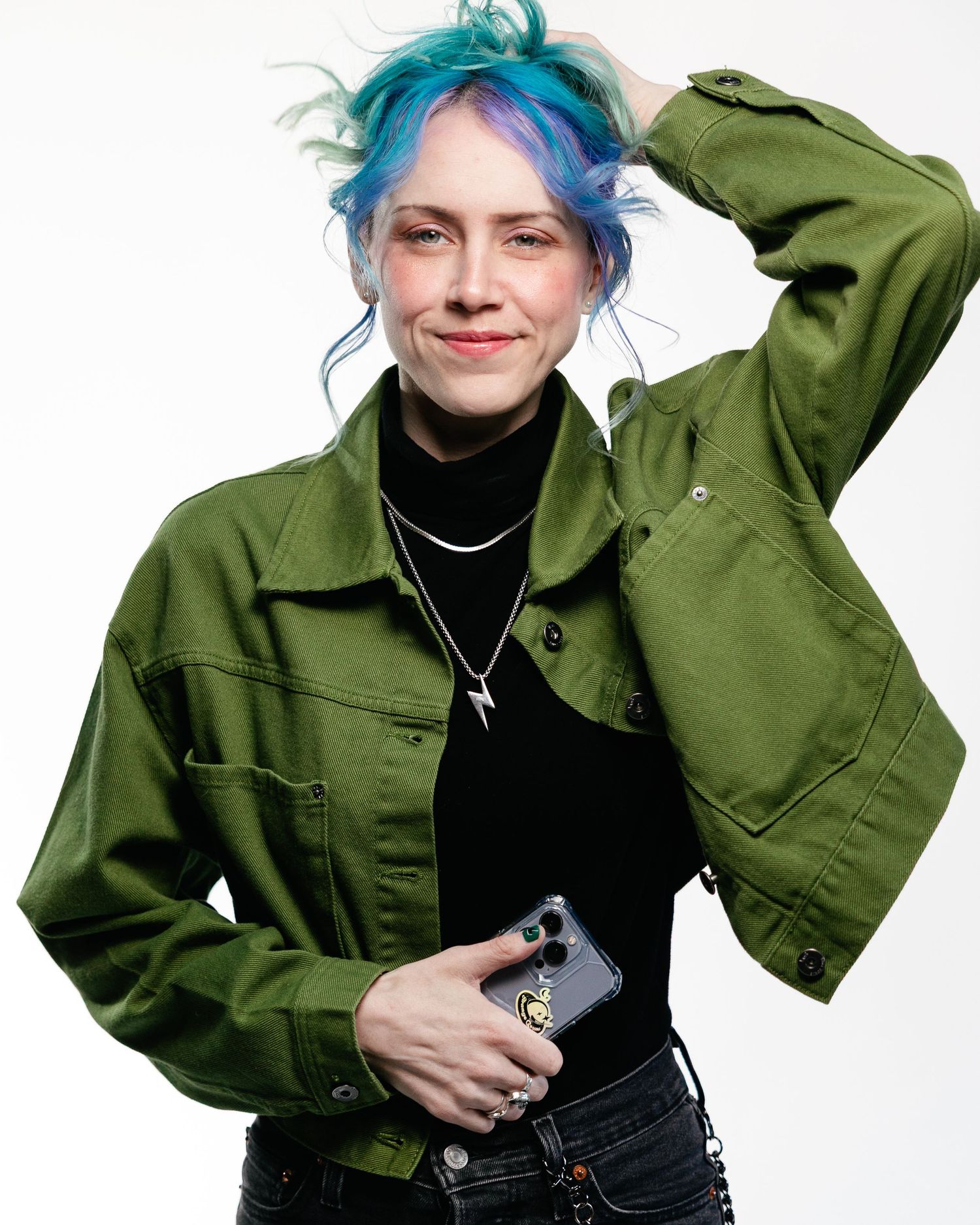 A person with vibrant blue hair poses in an olive green military-style jacket against a white background.