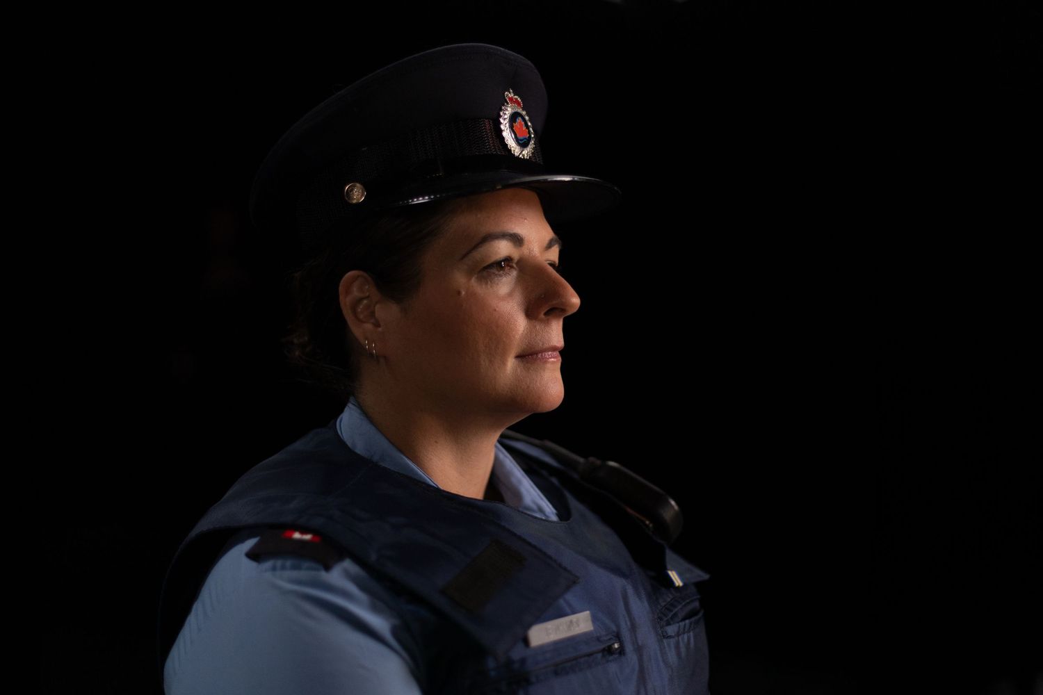 A police officer in dark uniform and cap stands in profile view against a black background with dramatic lighting.