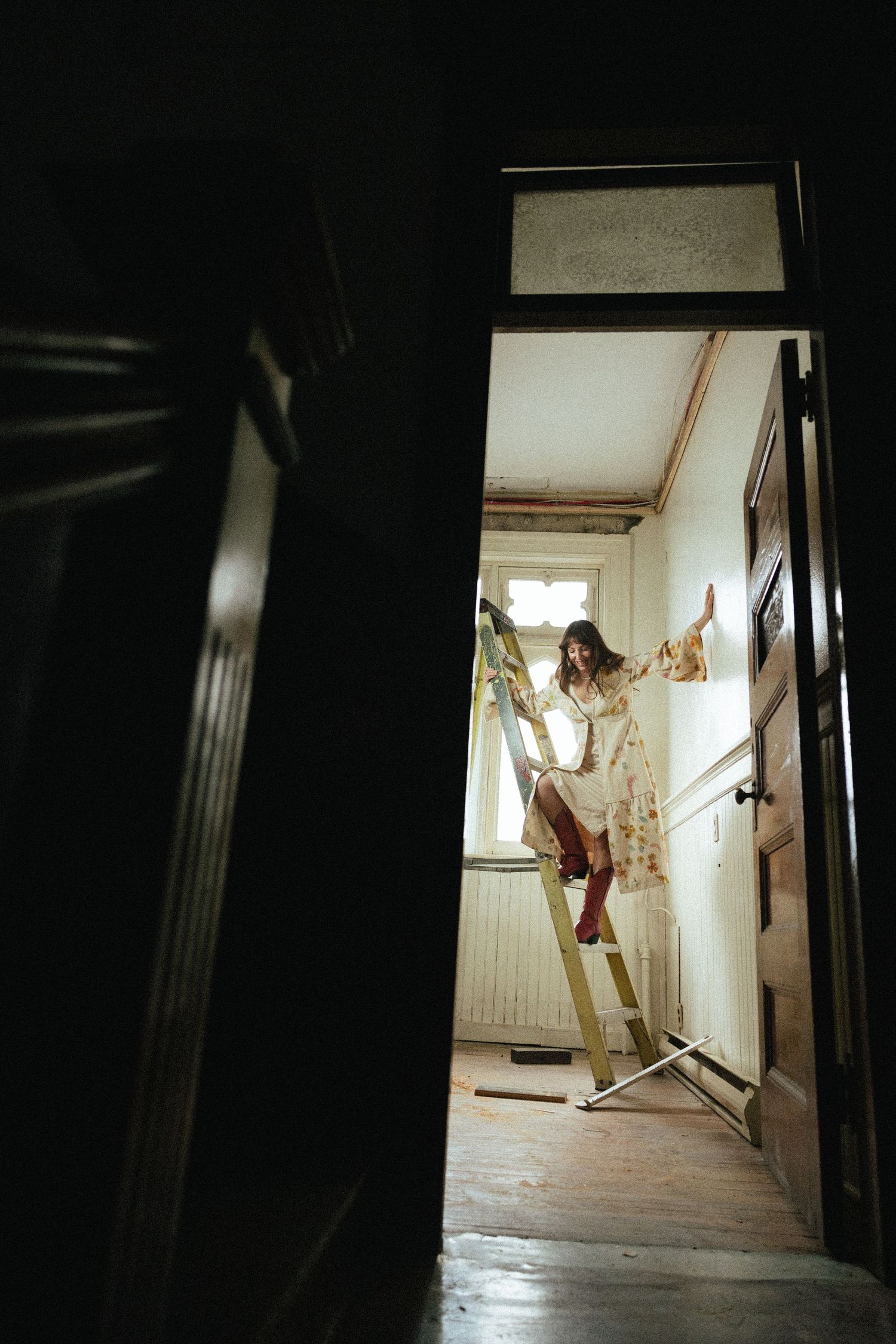 Through a dark doorway, a figure stands precariously balanced on a ladder in a dimly lit hallway.