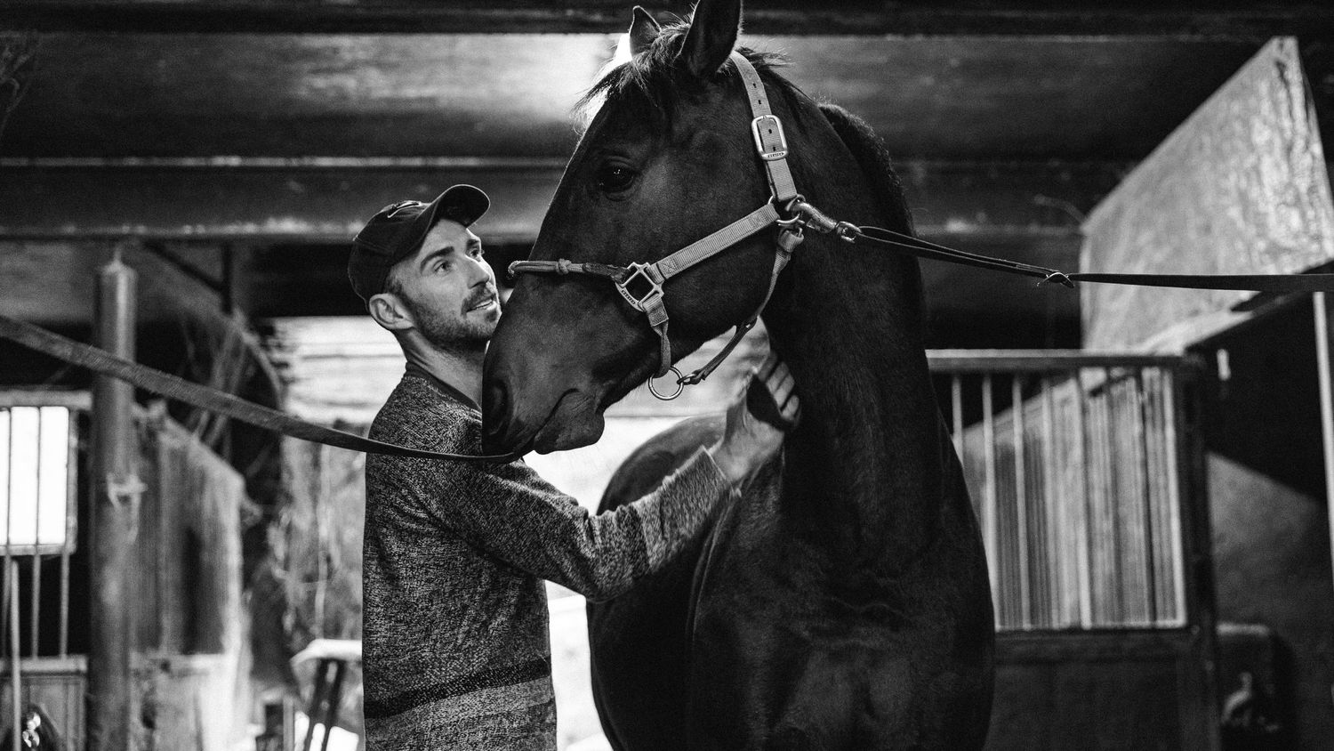 A black and white photograph of someone attentively grooming a dark horse in a wooden stable.