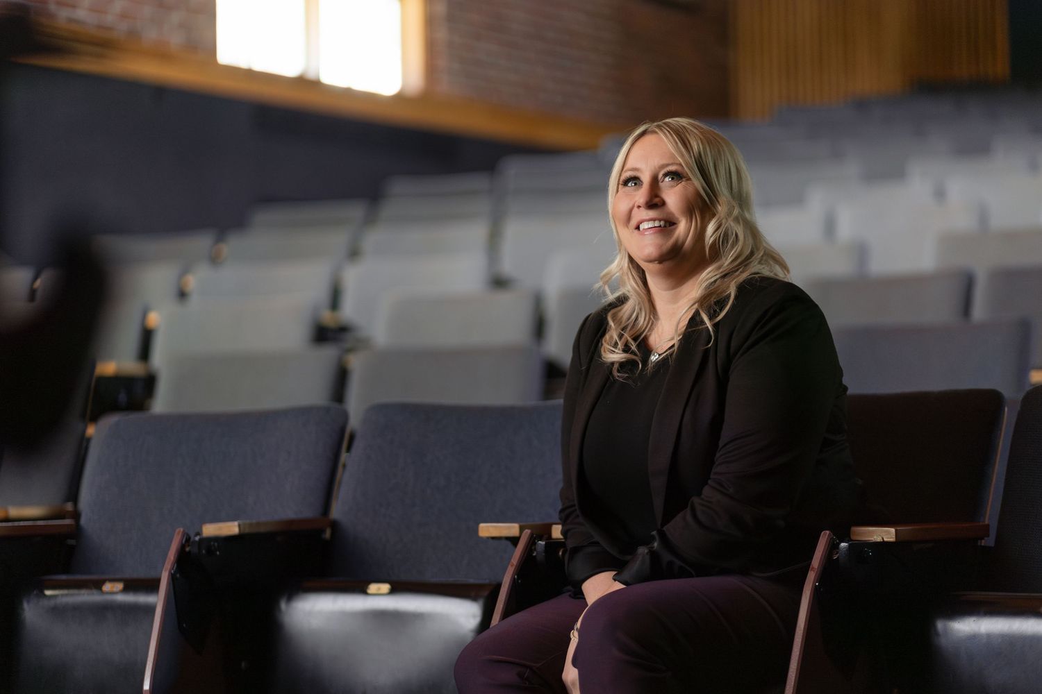 Person sits smiling in an empty theater auditorium with rows of navy blue seats and dramatic lighting.