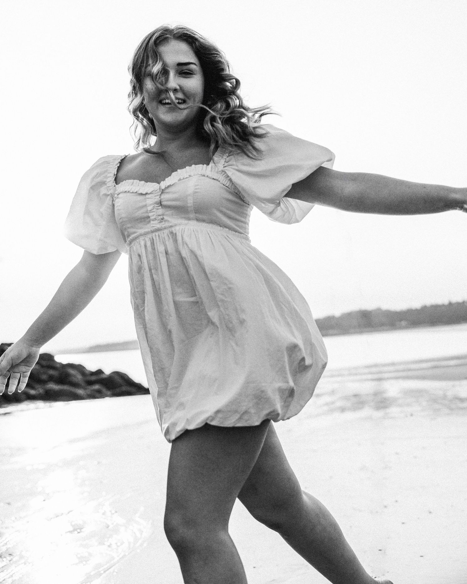 A joyful person in a white summer dress dances and twirls on a beach in black and white photography.