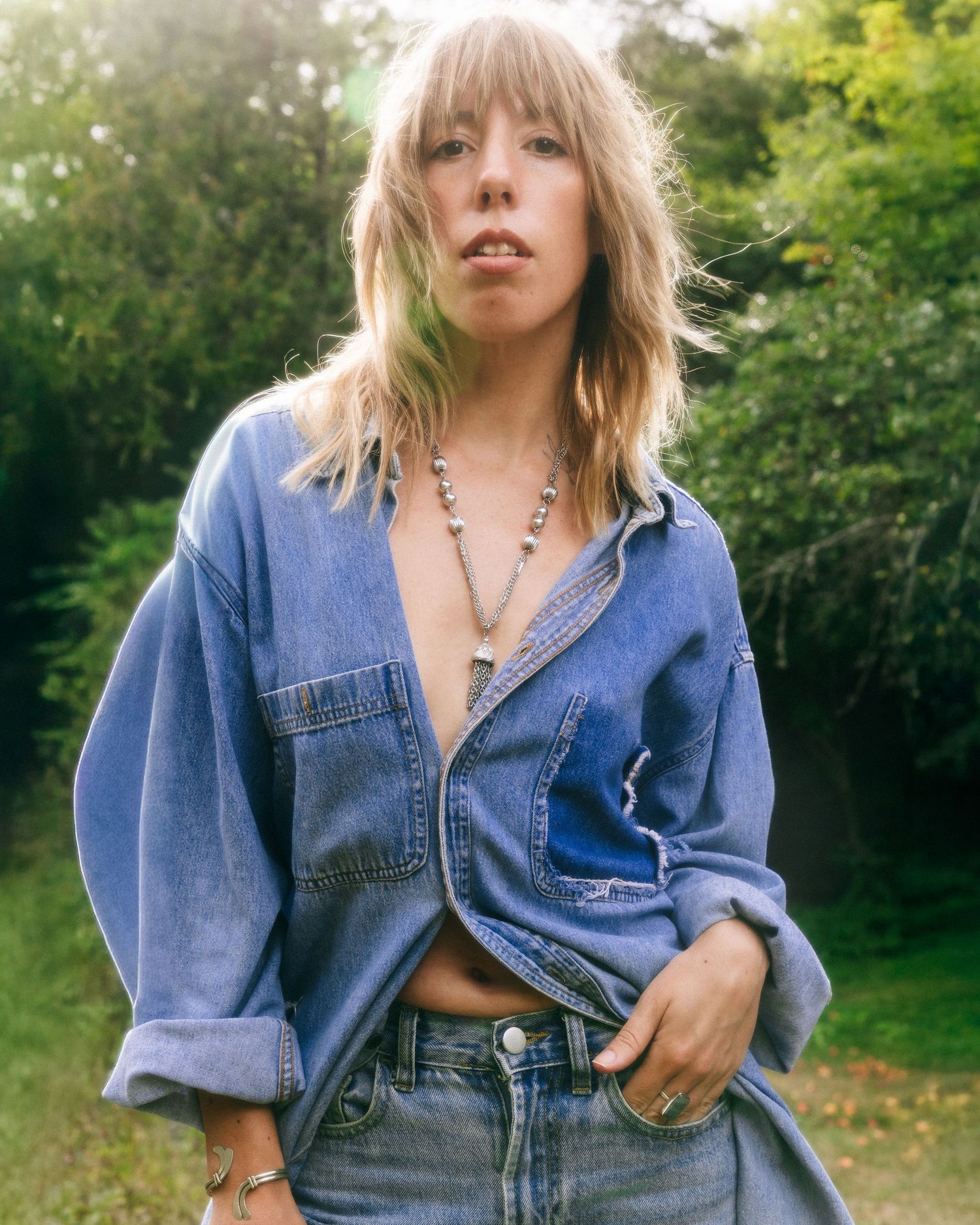 Person wearing denim jacket and jeans poses outdoors in natural sunlit setting with greenery in background.