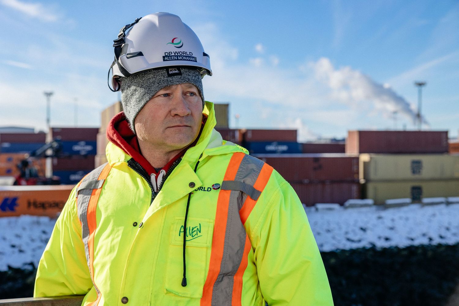Worker wearing high-visibility safety jacket and hard hat stands at industrial site in snowy conditions.