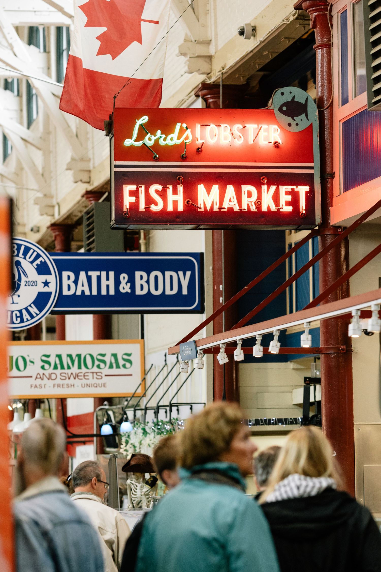 Busy indoor market hallway with storefront signs including Fish Market, Bath & Body and a Canadian flag.