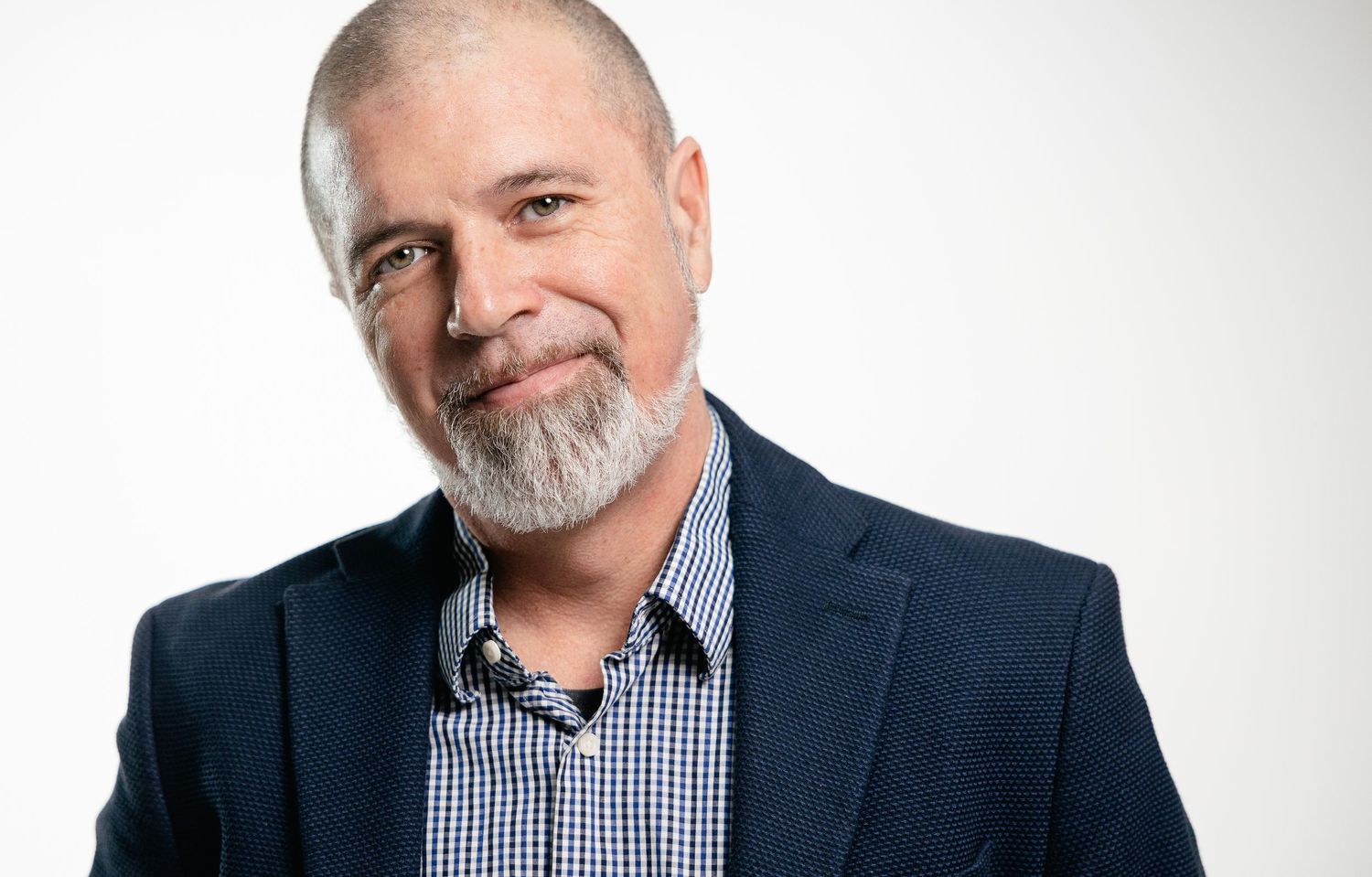 Professional headshot against white background showing person in navy blazer and checkered shirt with grey beard.