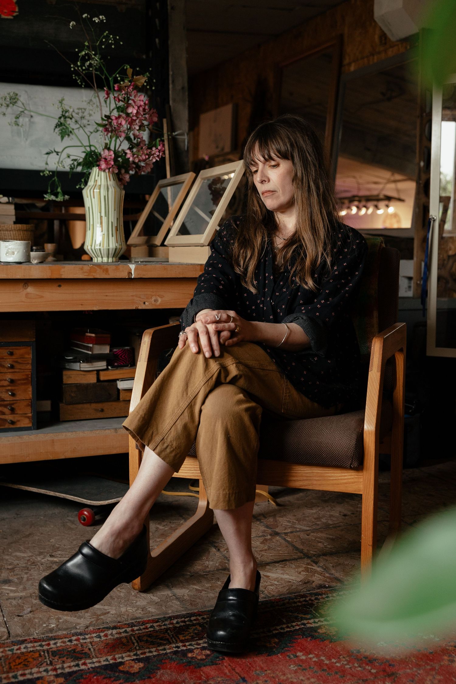 Figure in dark sequined top and brown pants sits thoughtfully in wooden chair at rustic table with floral arrangement.
