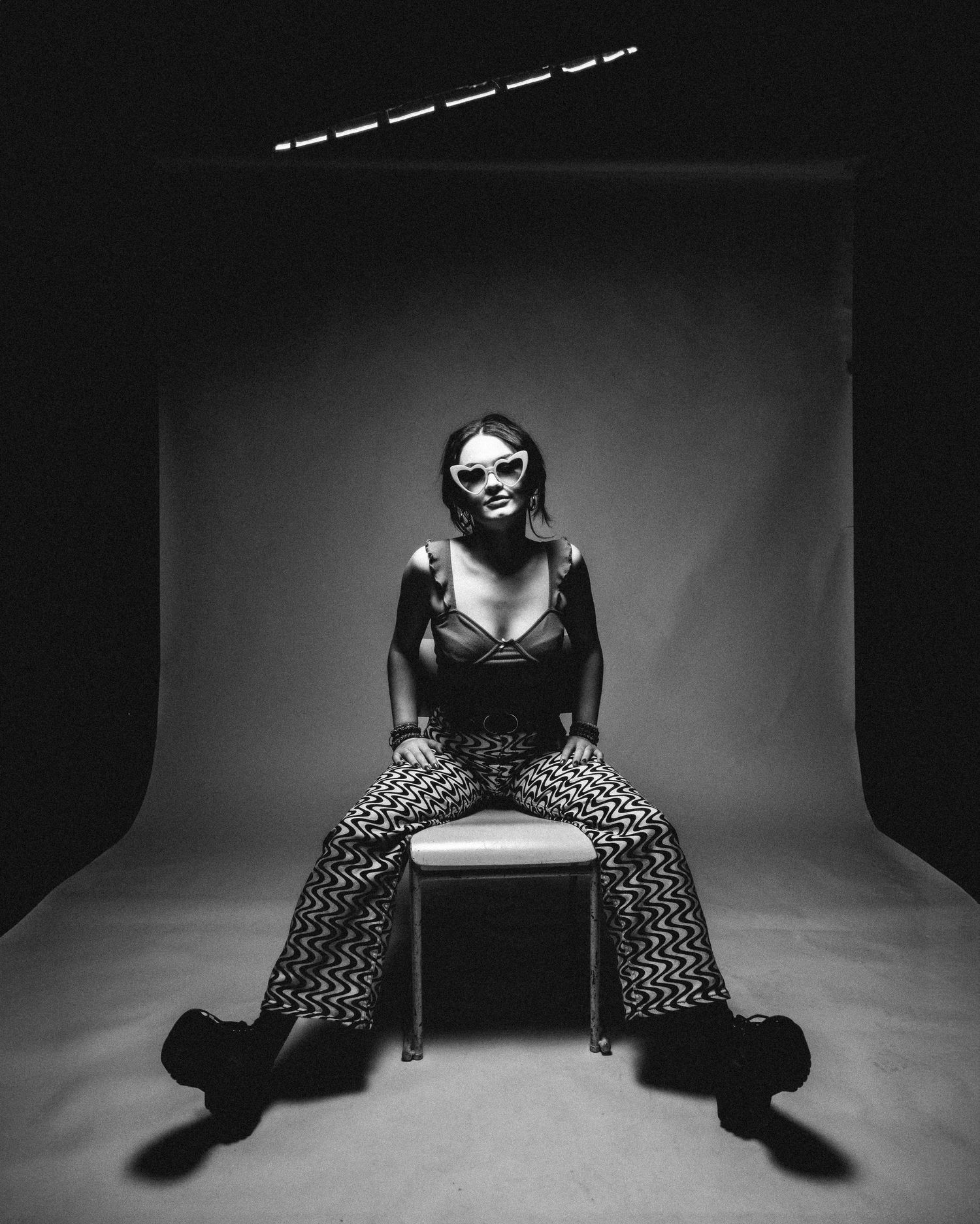 Black and white studio photograph showing a figure in patterned pants seated on a cube against a dark backdrop.