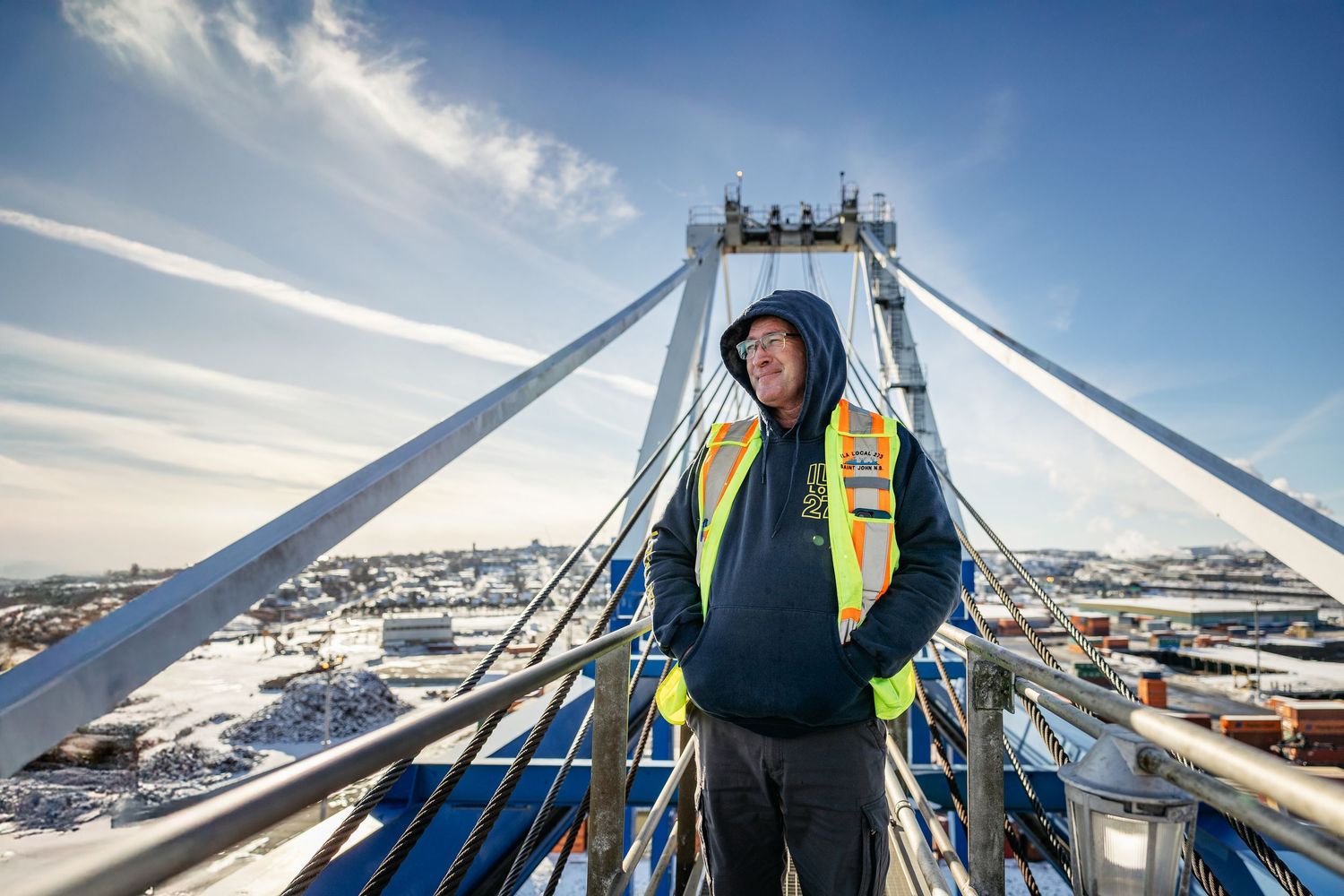 Worker in safety gear stands high up on a snowy bridge structure with bright blue sky and winter cityscape in background.