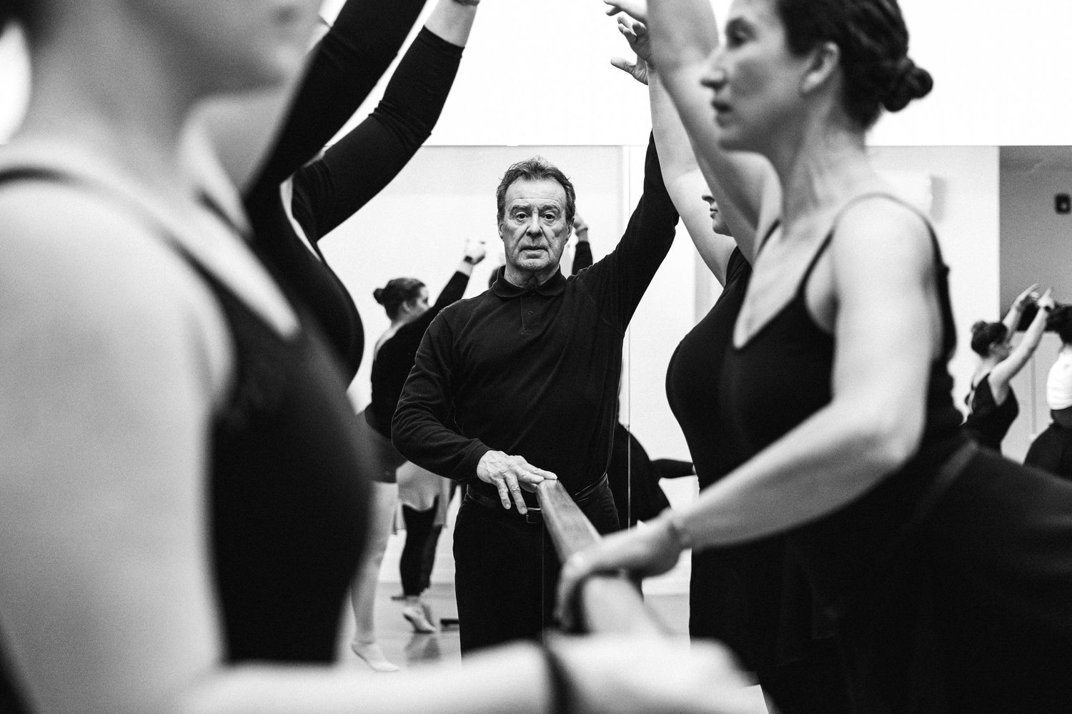 A ballet instructor leads a dance class, gesturing enthusiastically as dancers in black leotards practice in the studio.
