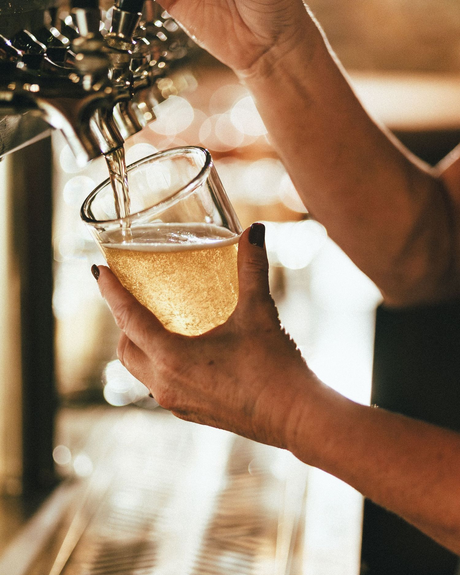 Golden beer being poured from a tap into a glass at a bar with warm lighting in the background.