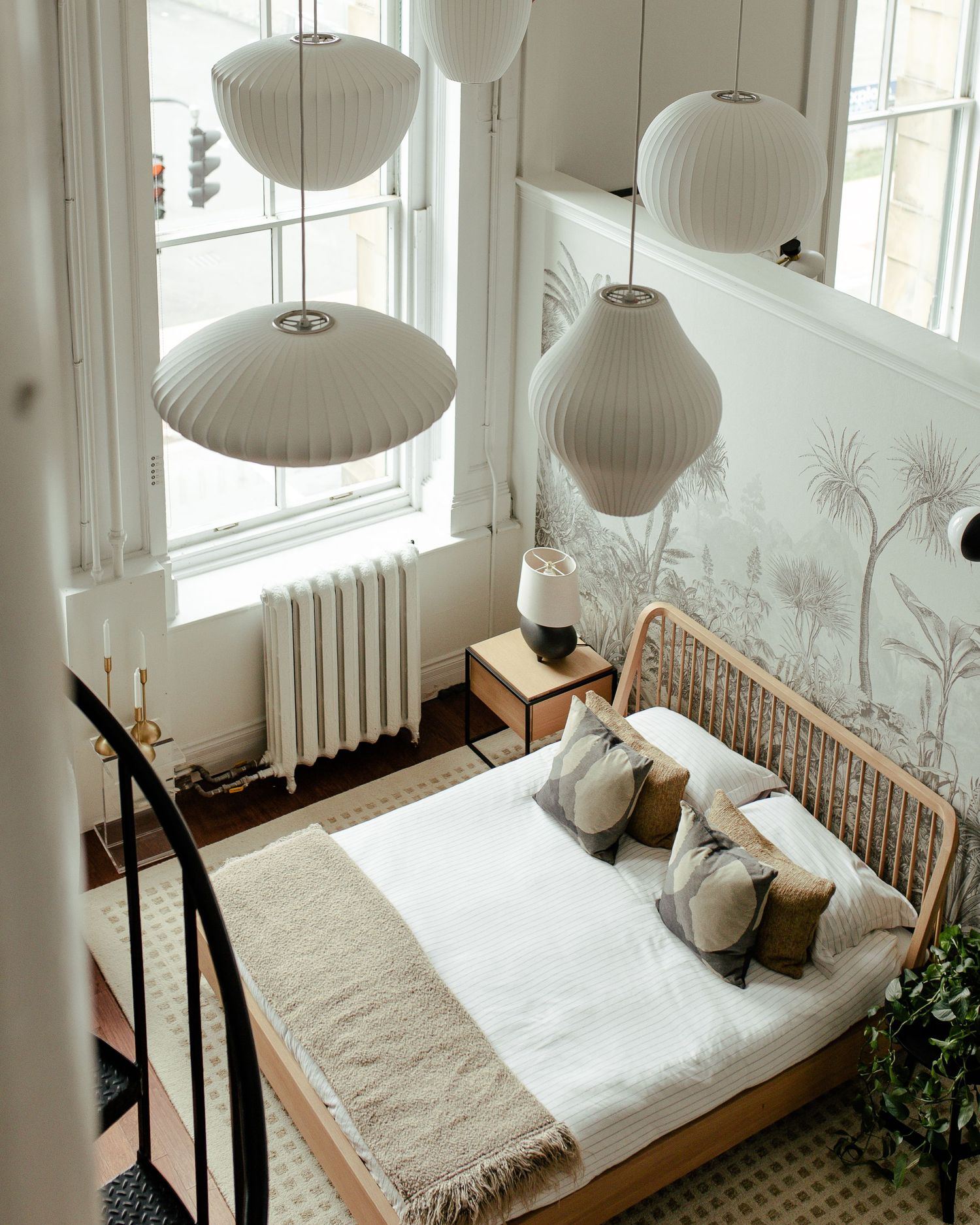 Cozy bedroom corner with white pendant lamps, rattan bed frame, linen bedding and botanical wallpaper accents.