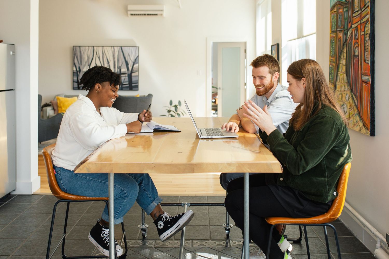 Three colleagues collaborating at wooden table in bright office space with wall art and natural light.