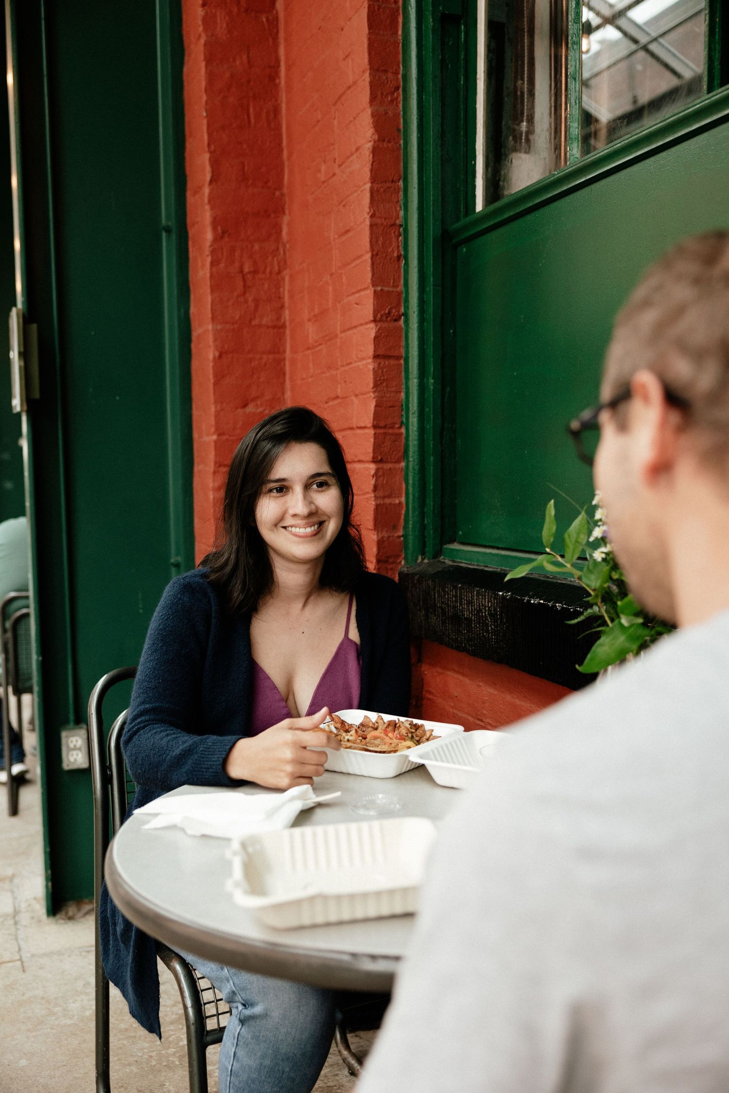 Two people sitting at an outdoor cafe table enjoying a casual meal on a patio with brick and green painted walls.