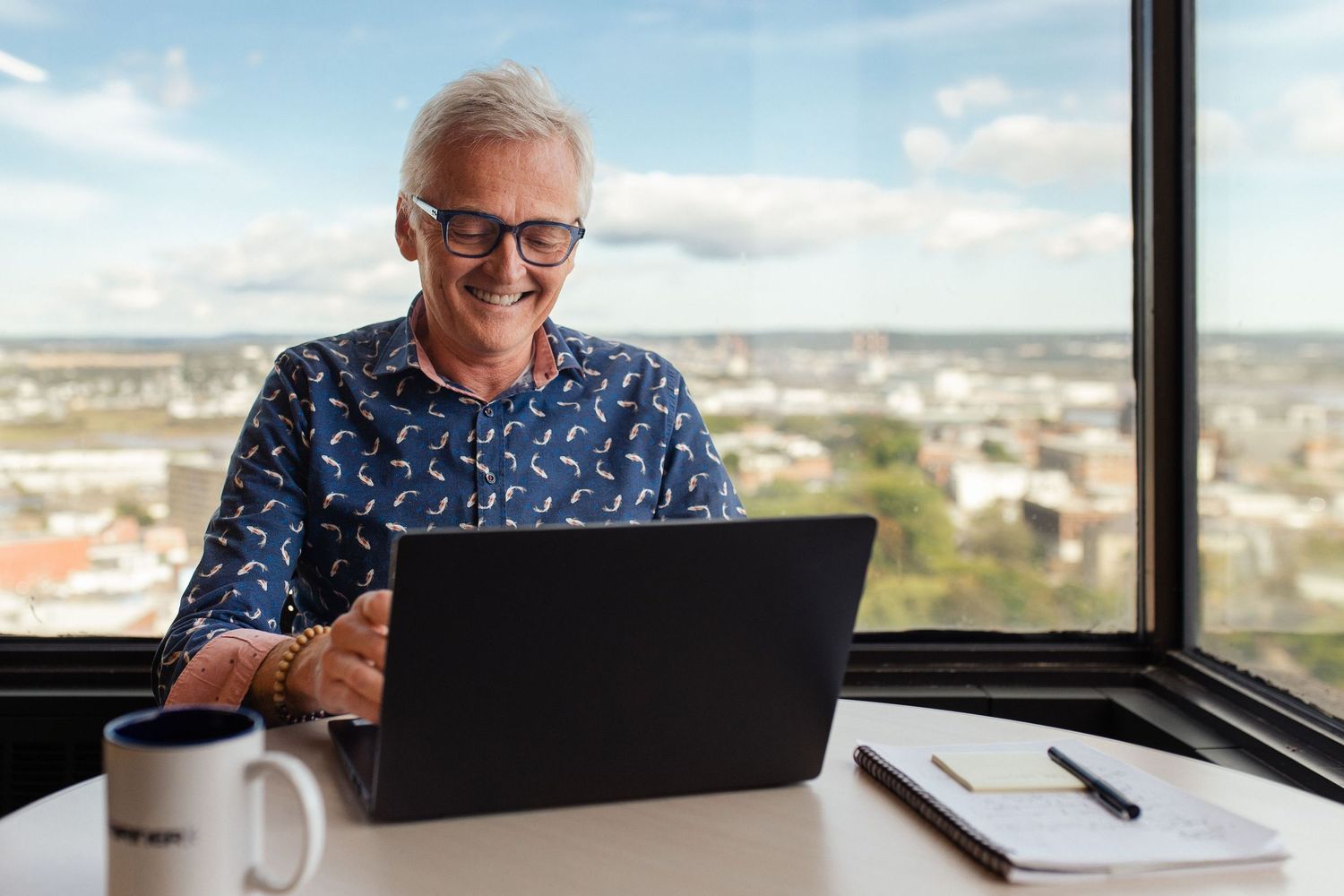 Person in a floral shirt working on a laptop computer at a desk with a city view in the background.