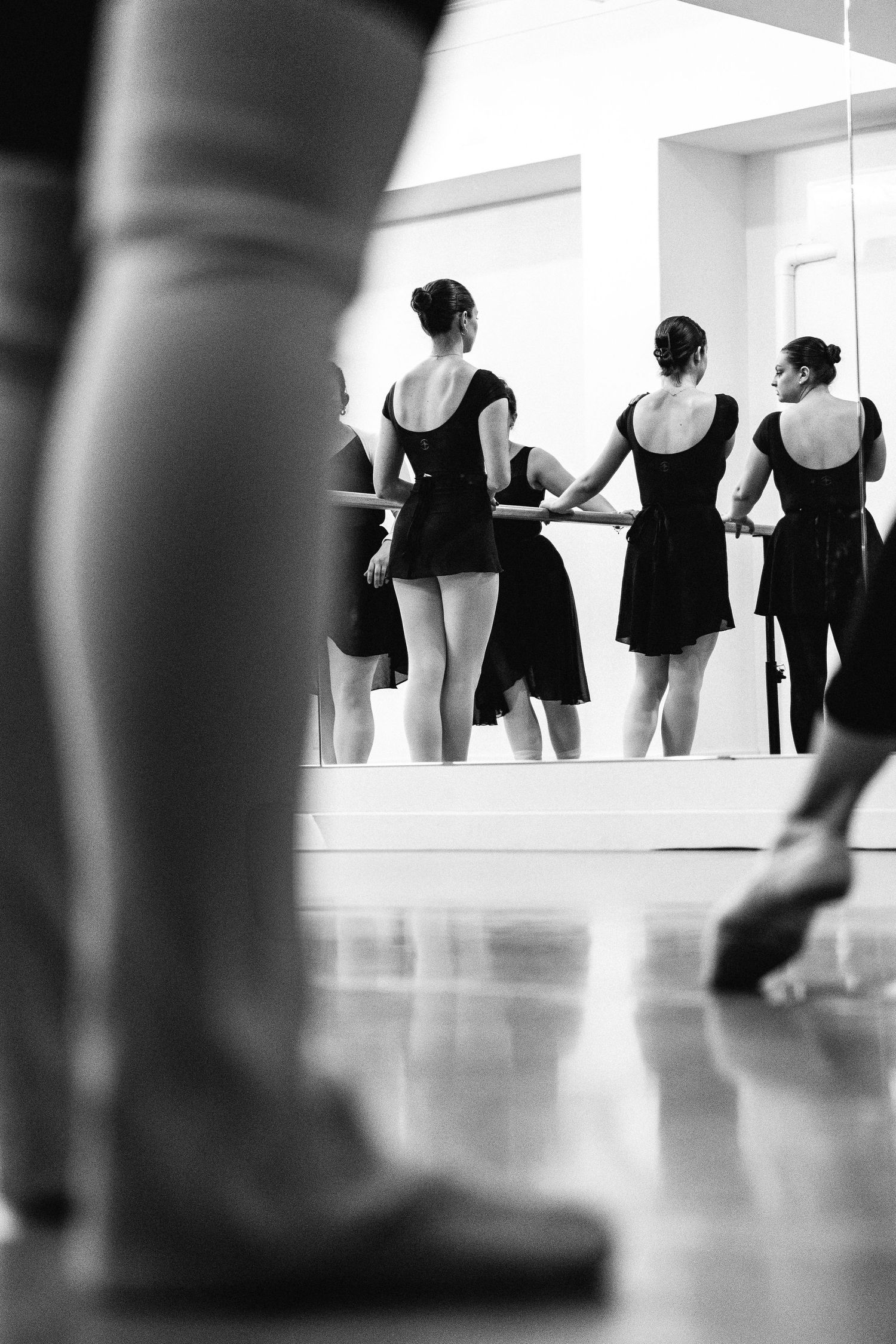 Black and white image of dancers in leotards practicing at a ballet studio with a mirror reflection visible.