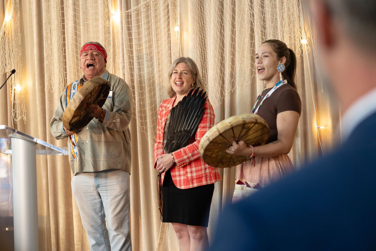 Three musicians perform together on stage with drums and percussion instruments while sharing joyful laughter.